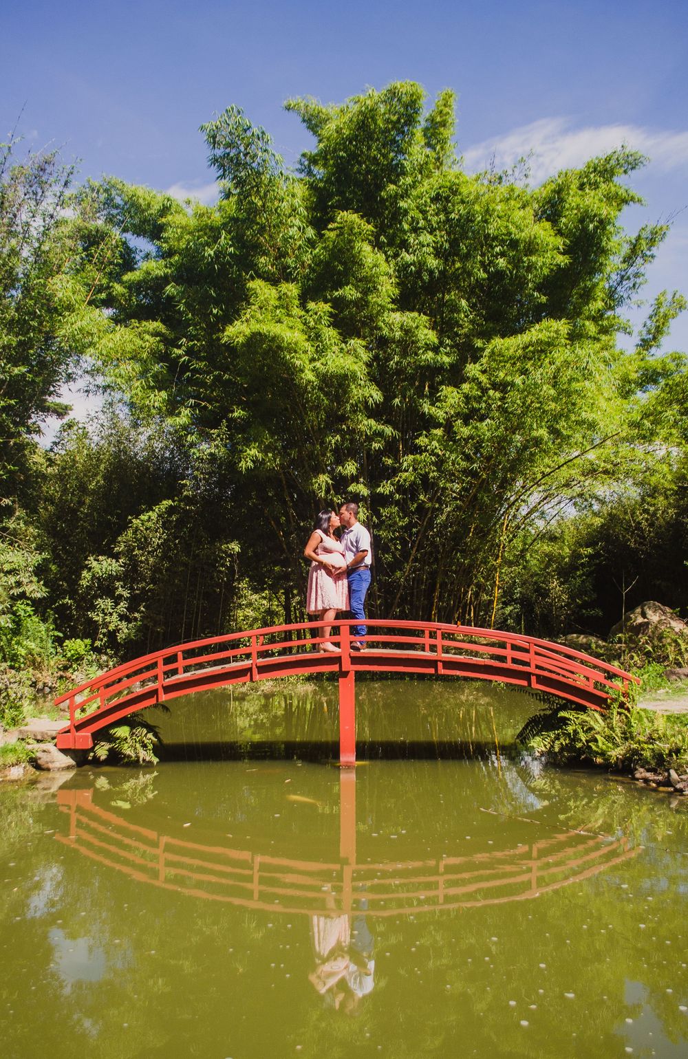 Couple embracing on a red arched bridge over a serene pond, surrounded by lush green bamboo trees.