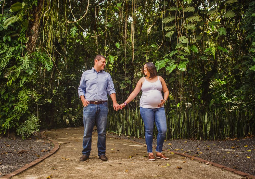 Pregnant couple holding hands on a garden path surrounded by lush tropical greenery during a maternity photo shoot.