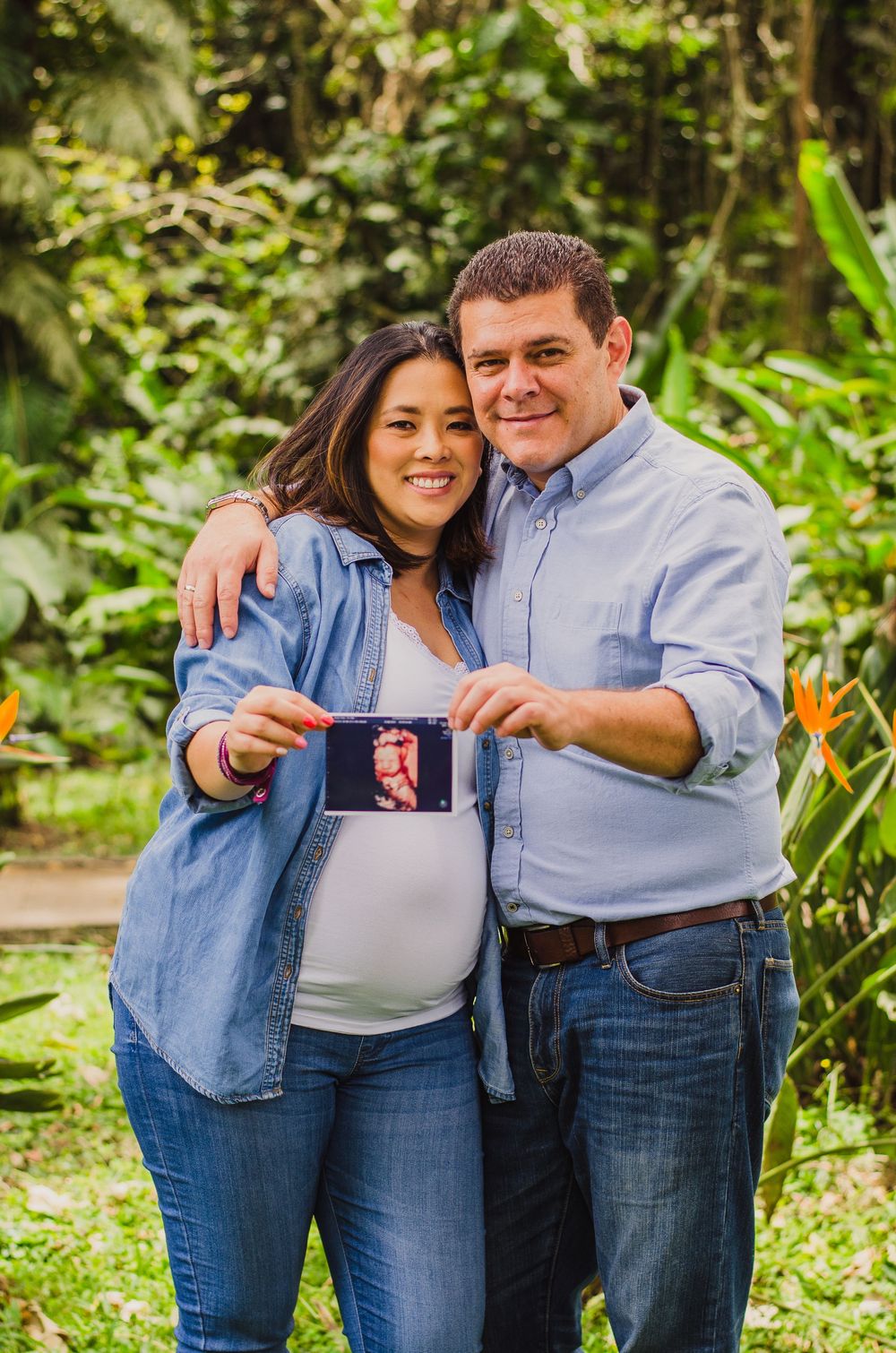 Smiling couple holding ultrasound photo outdoors, pregnant woman in denim shirt with partner in lush green garden setting.