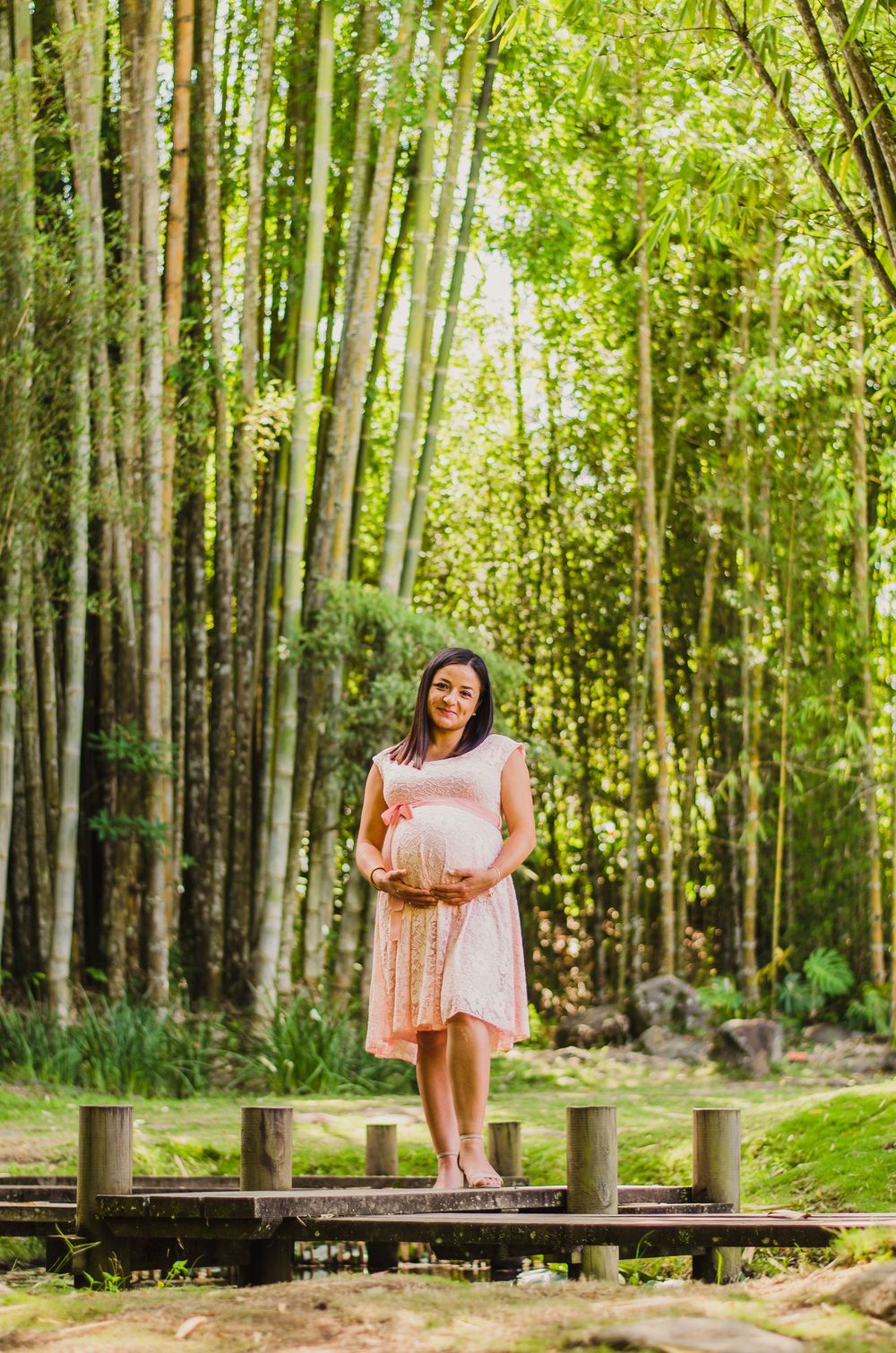 Pregnant woman in pink dress cradling baby bump on wooden bridge in lush bamboo forest.