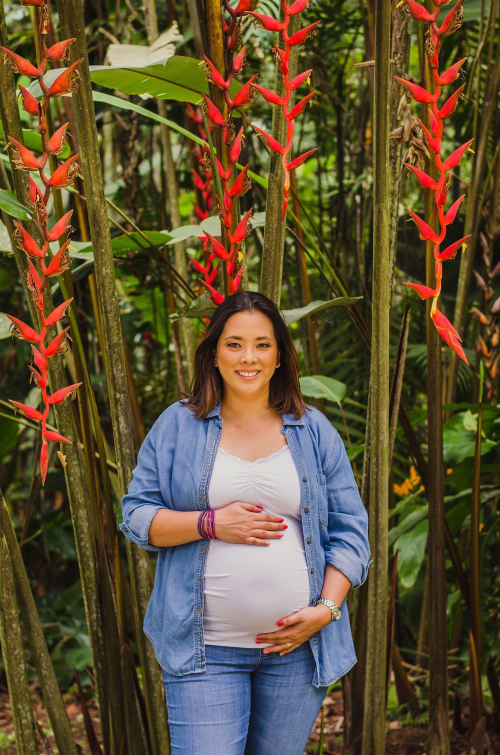 Smiling pregnant woman in denim shirt holding baby bump surrounded by tropical red heliconia flowers.
