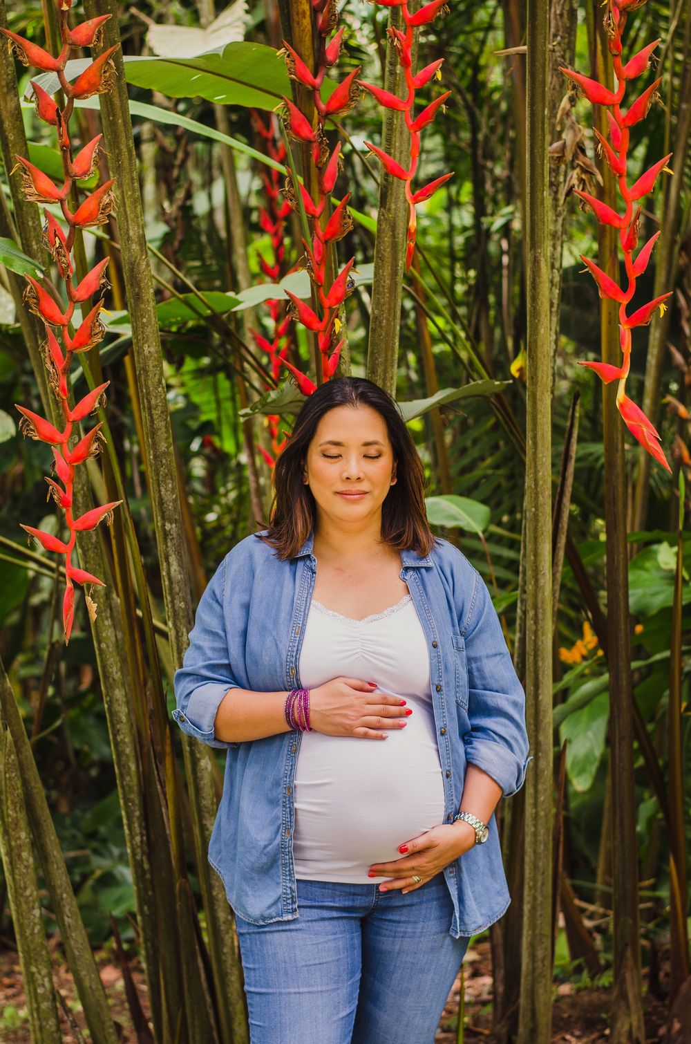 Pregnant woman in denim shirt cradles baby bump outdoors among tropical red heliconia flowers and green foliage.
