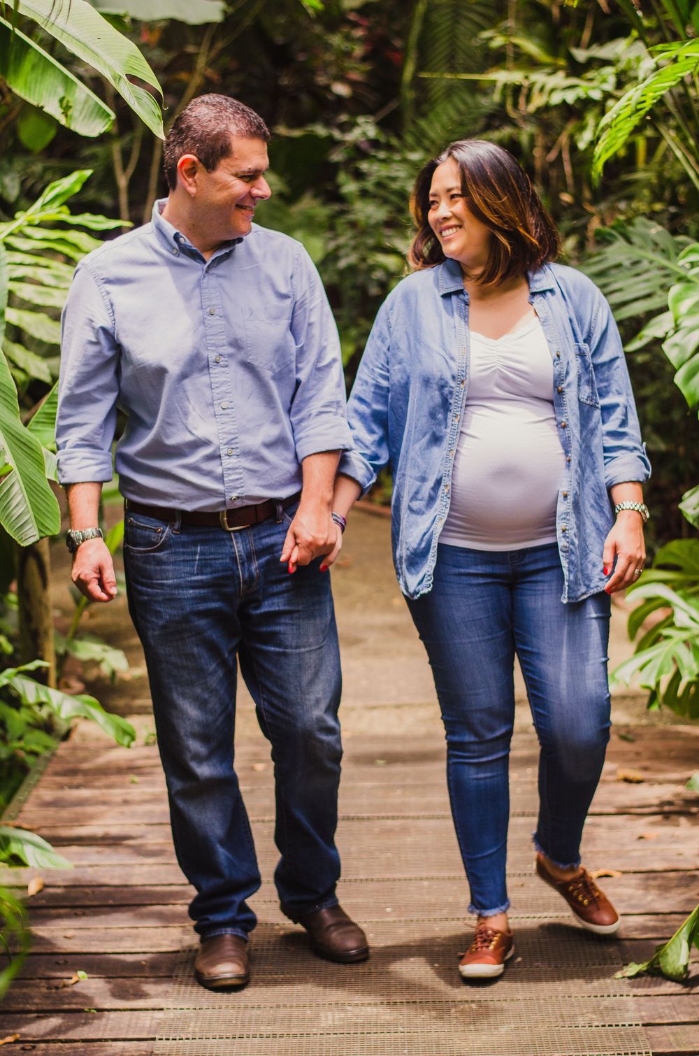 Expecting couple holding hands and smiling while walking on a wooden path surrounded by lush tropical greenery.