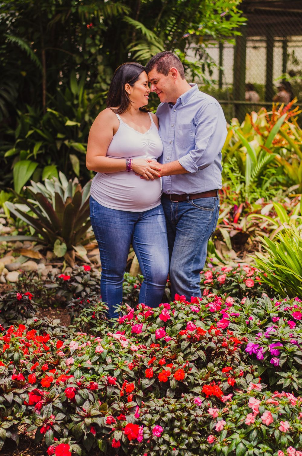 Pregnant couple embracing tenderly in a lush garden surrounded by vibrant red and pink flowers during a maternity photoshoot.