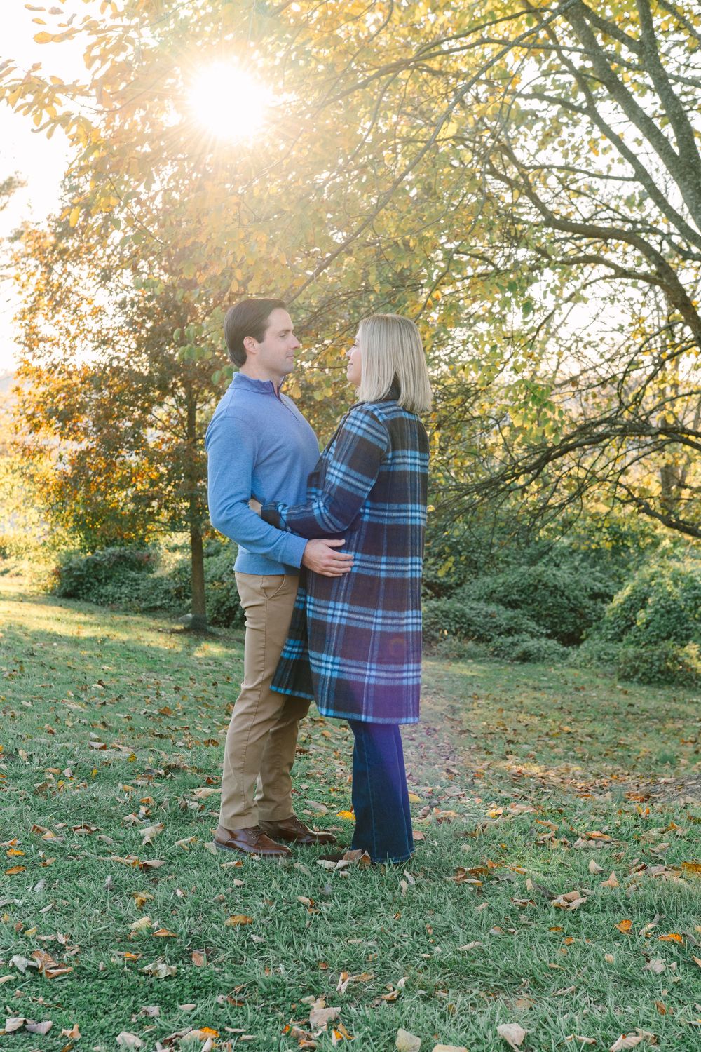 Couple standing together on grass with golden sunlight filtering through autumn trees.