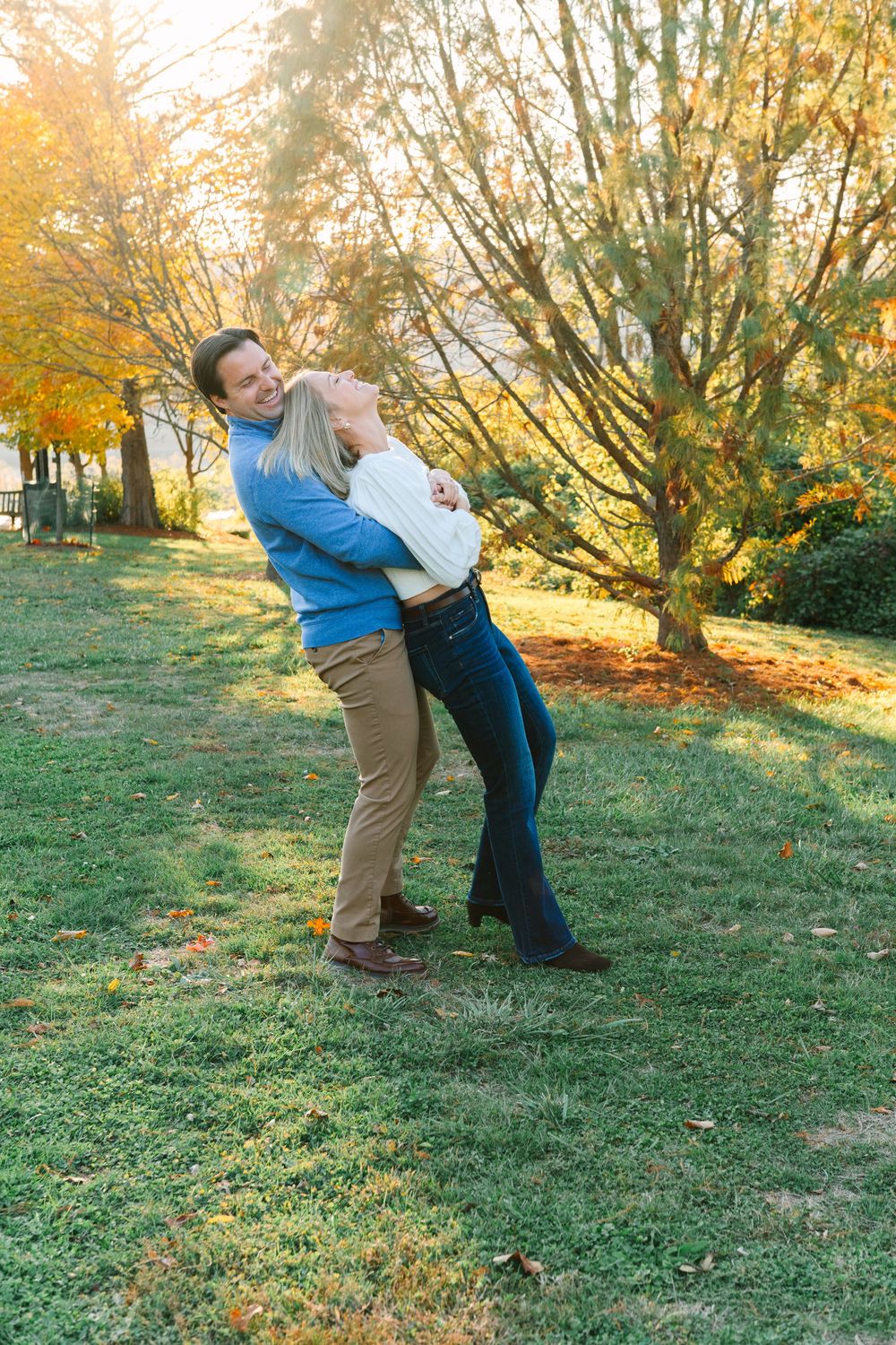 A couple shares a playful moment in a park with autumn trees in the background.