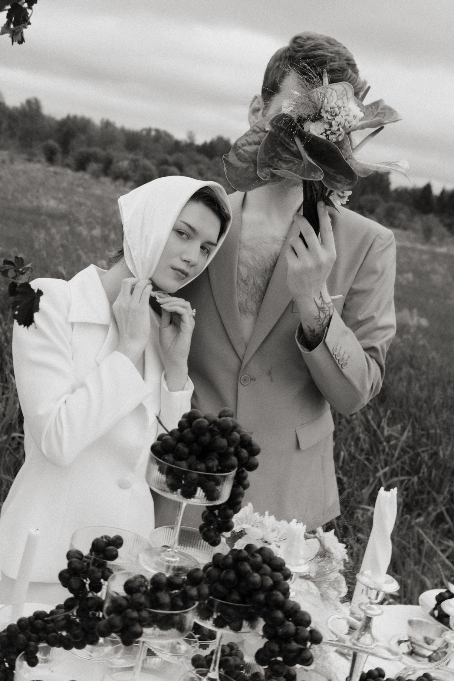Black and white photo of a bride's flowing white dress train and groom's legs in a grassy meadow.