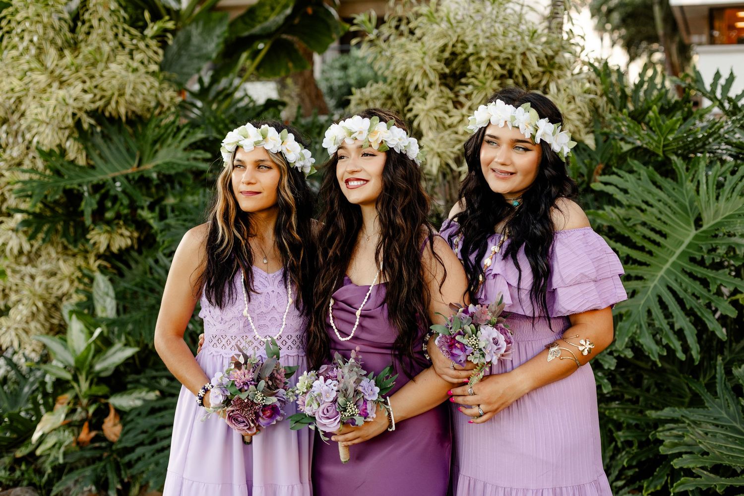 Three bridesmaids in lavender dresses with flower crowns pose with bouquets against tropical foliage.
