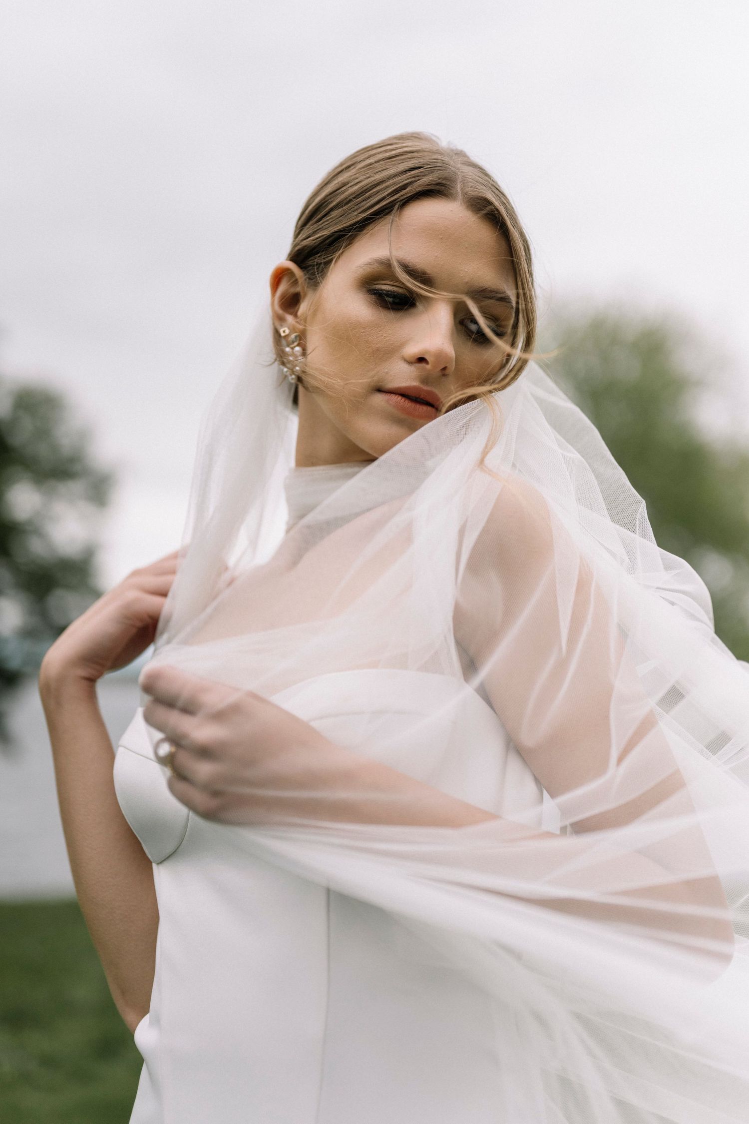 Ethereal bridal portrait featuring delicate sheer veil flowing in breeze against overcast sky and natural backdrop at Glen Manor House in RI