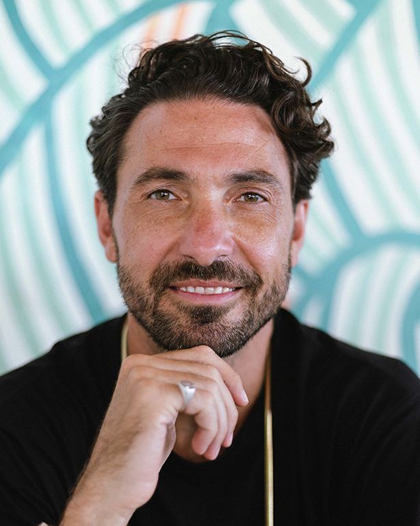 Professional headshot of a stylish man with beard against blue patterned background.