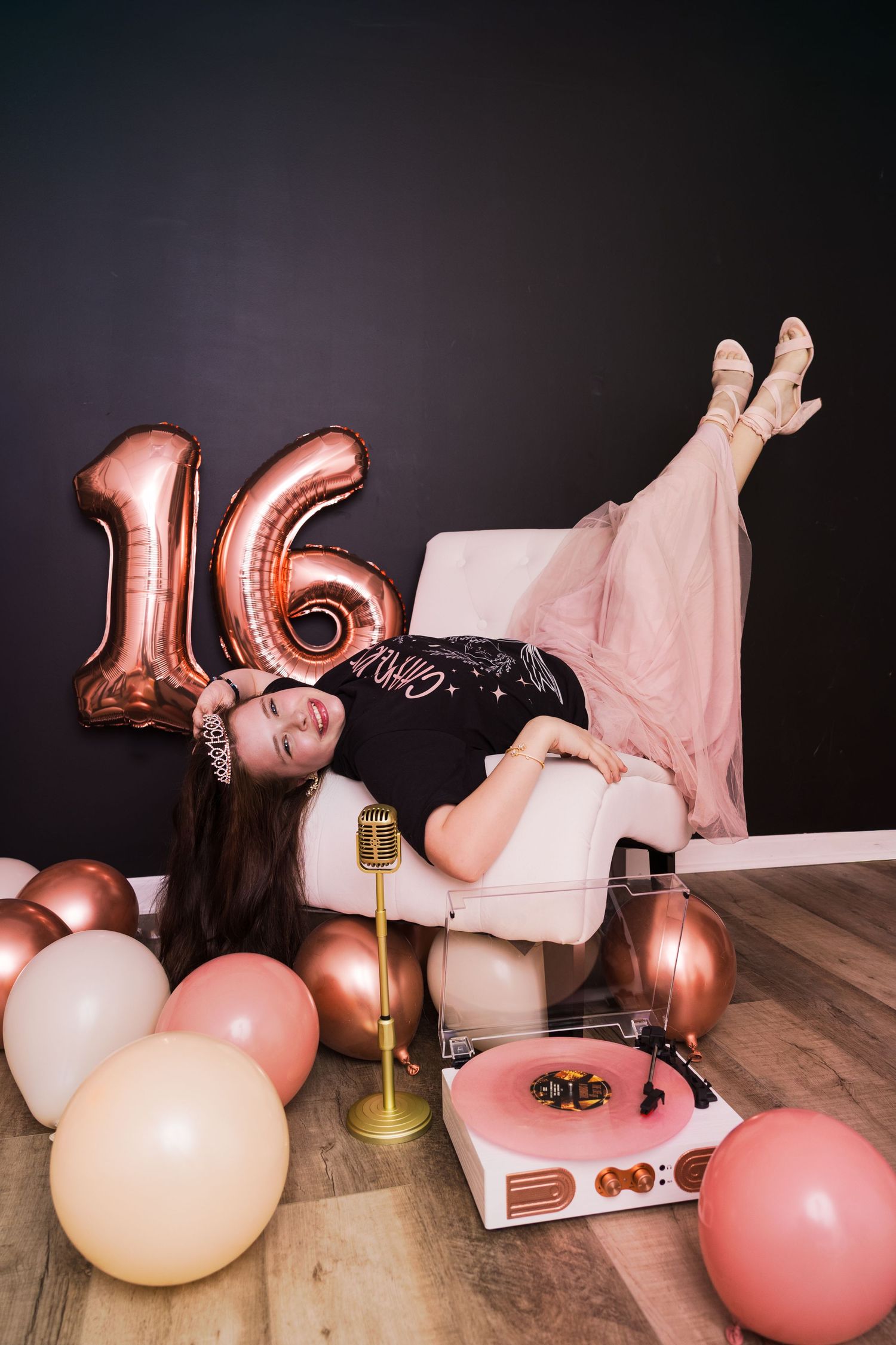 Teen girl lounging in white chair celebrating sweet 16 birthday with rose gold balloons and vintage record player.