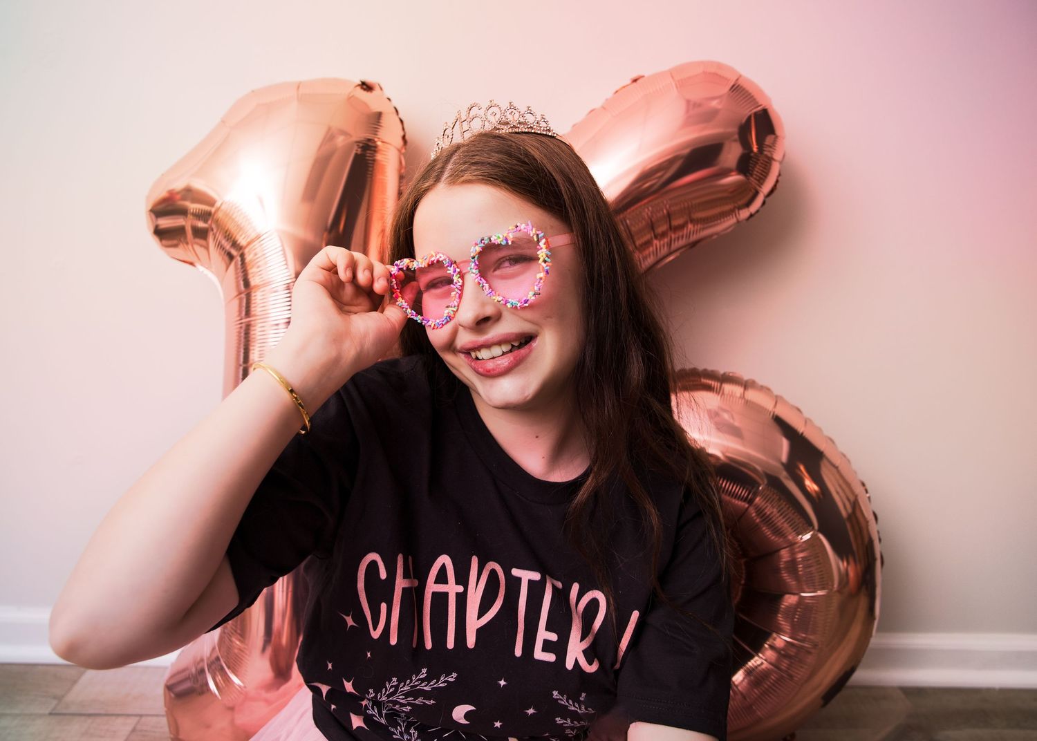 Happy teenage girl celebrating birthday with rose gold number balloons, sparkly tiara, and heart-shaped party glasses.