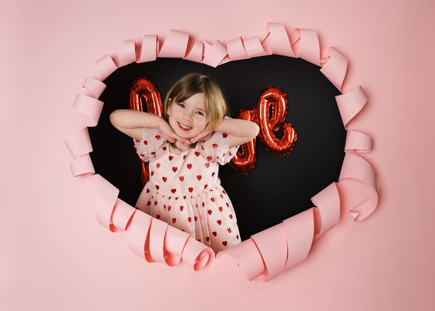 Smiling young girl peeking through torn pink heart-shaped paper backdrop for Valentine's Day themed photo session.