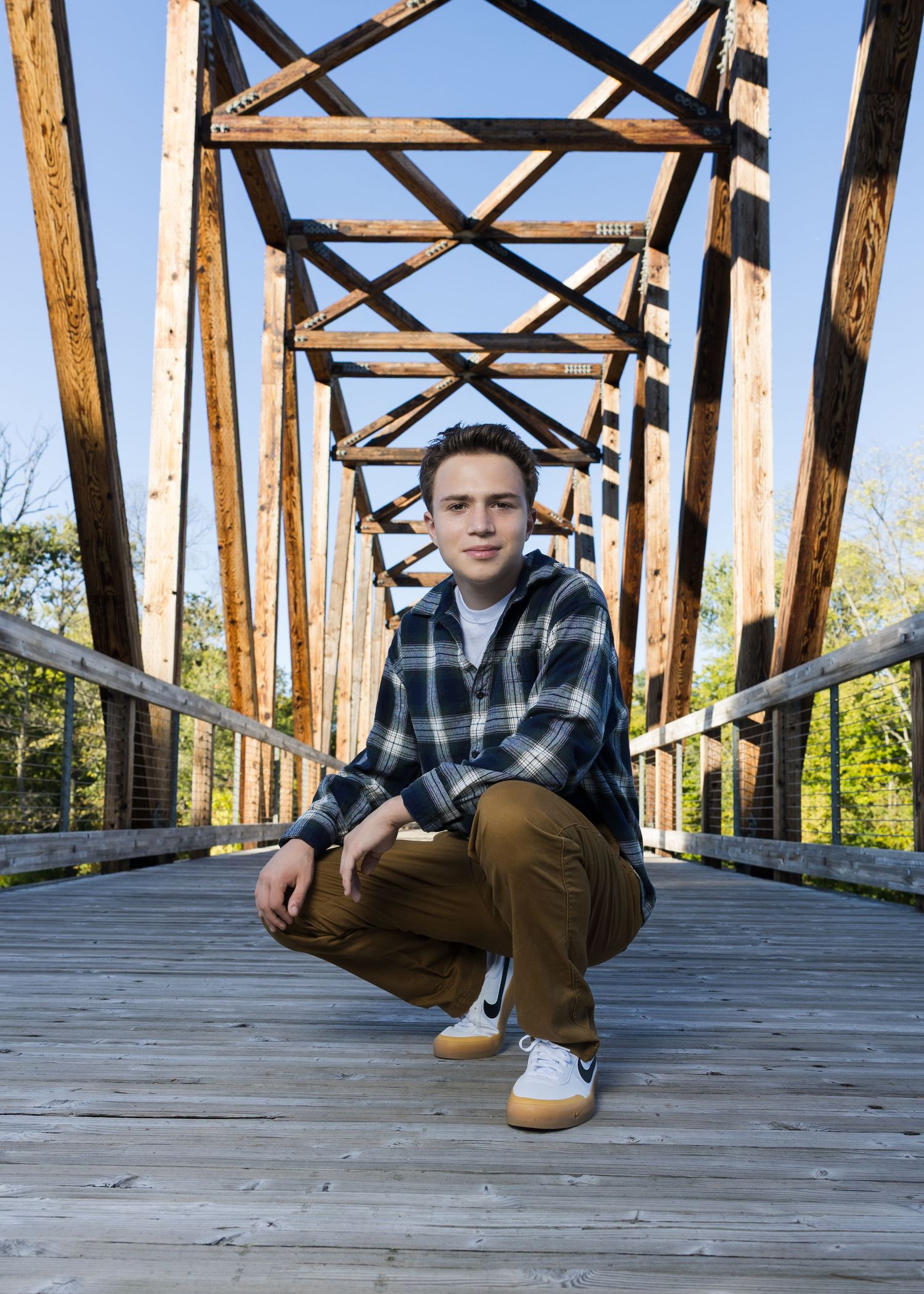 Teen boy in plaid flannel shirt crouching on wooden bridge for outdoor senior portrait photography session.