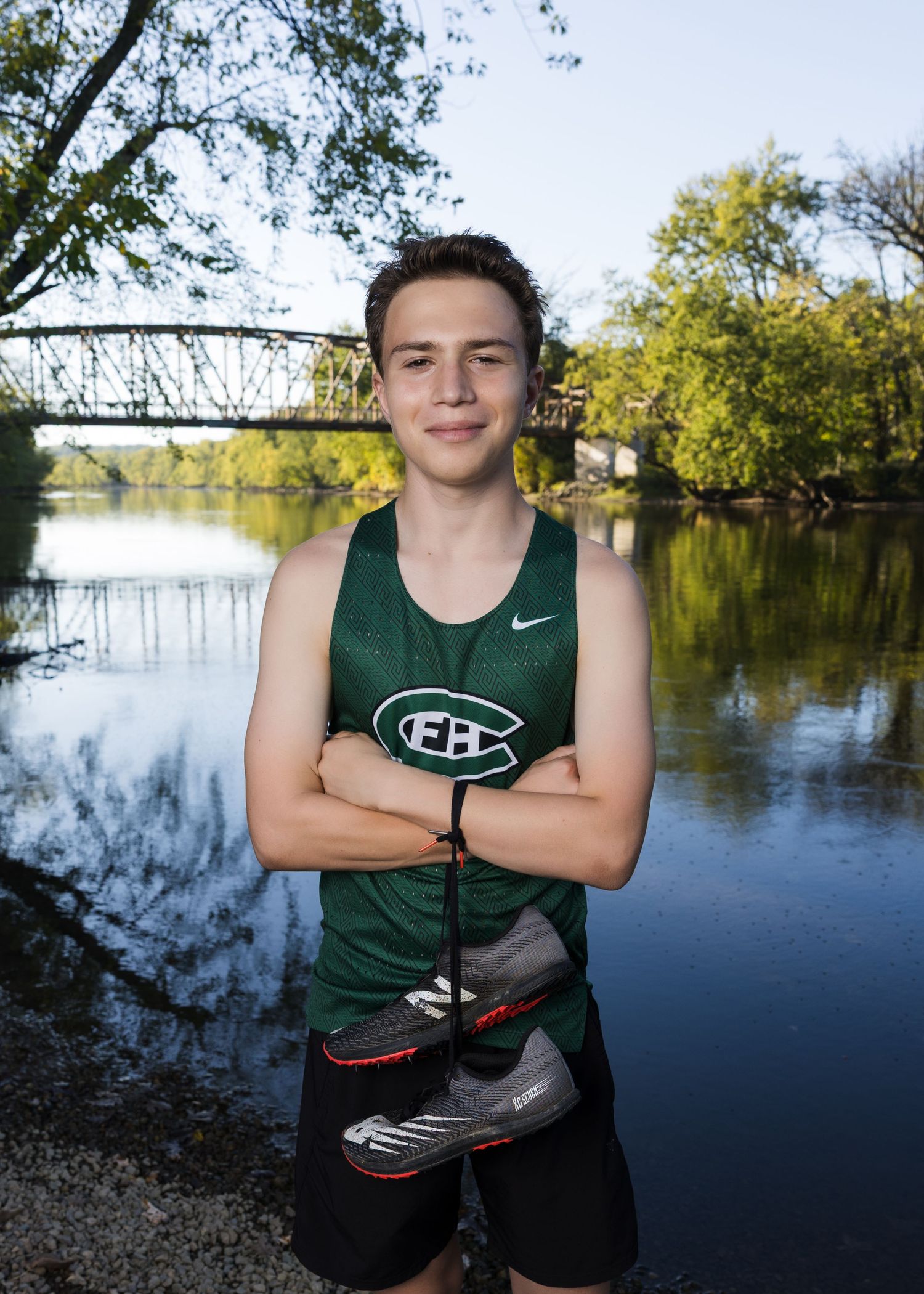 Young male athlete in green Nike cross country uniform posing with shoes by river and historic bridge backdrop.