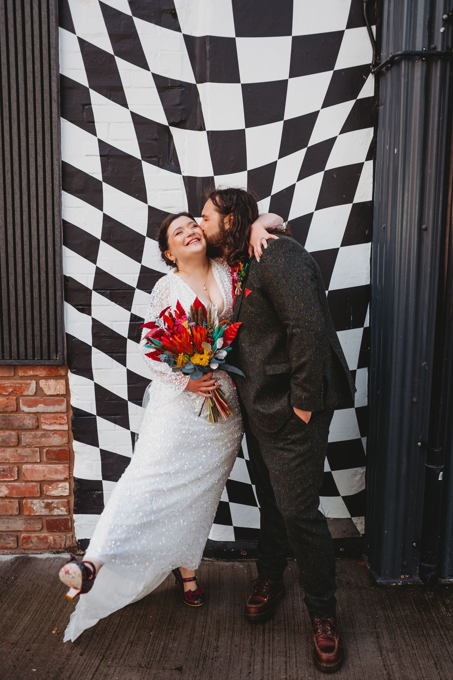 Bride and groom pose before a black and white checkered backdrop, kissing while holding a vibrant bouquet at Shack Revolution Hereford