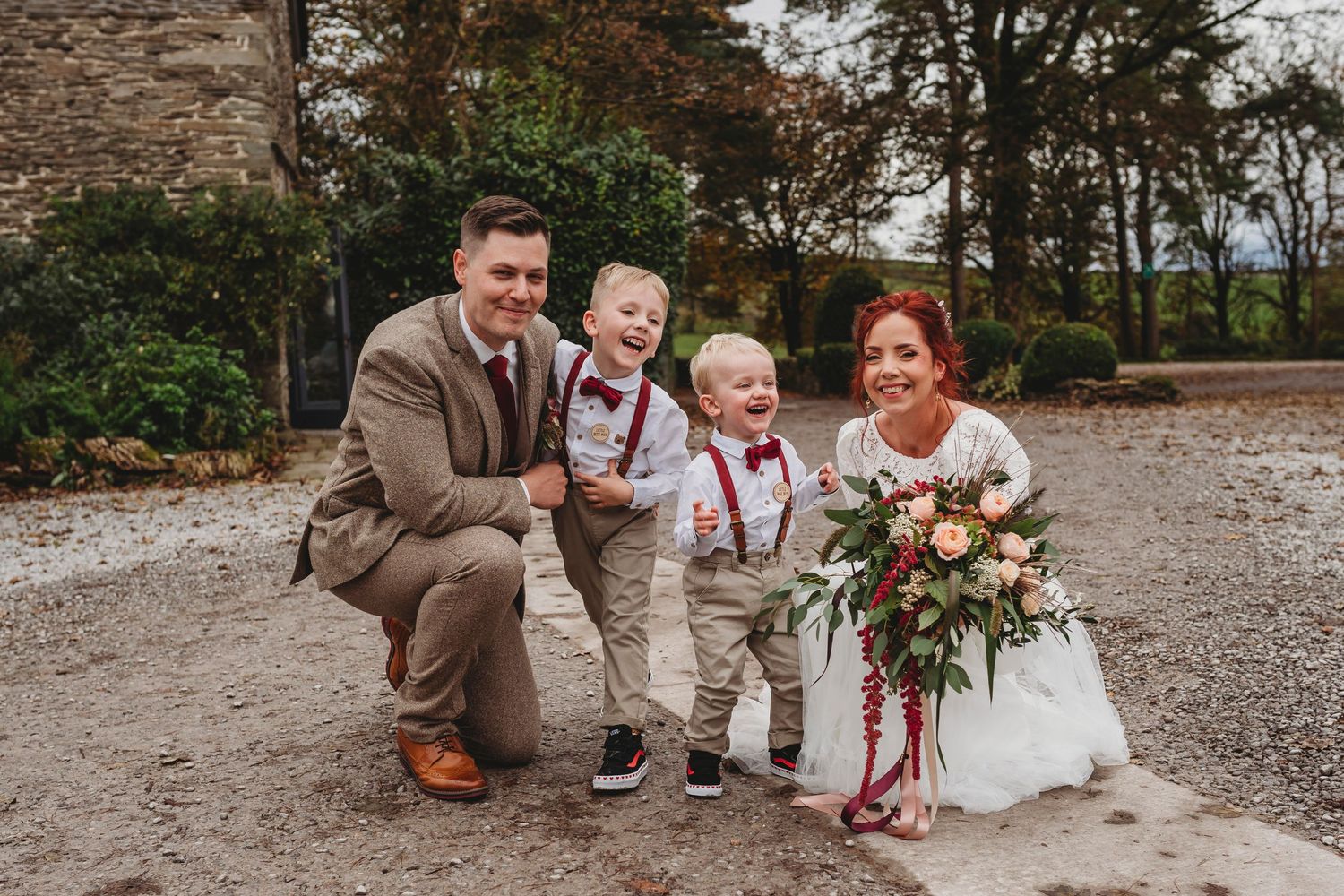 Wedding party poses outdoors on rustic gravel path with autumn foliage in background.