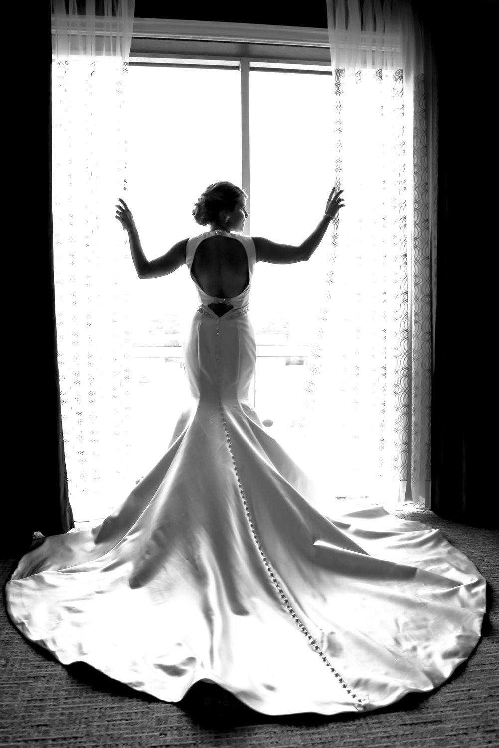 Black and white silhouette of bride in elegant wedding gown with long train poses gracefully by window with sheer curtains.