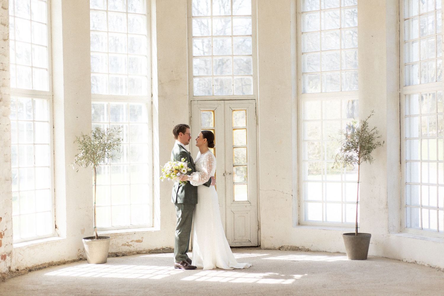 A sunlit wedding portrait in a bright white room with tall windows and potted plants.