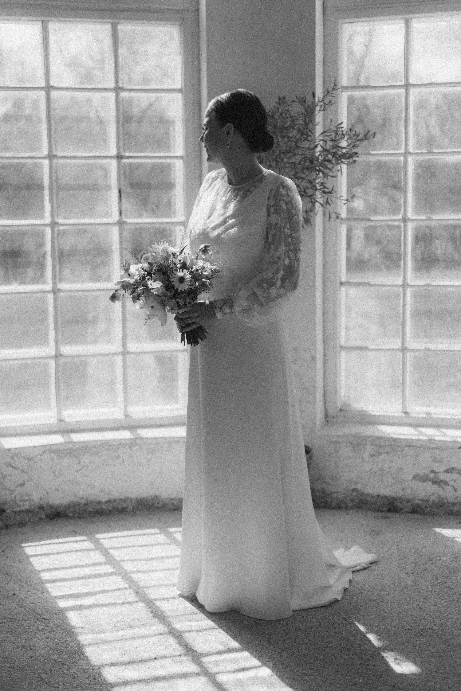 Elegant bride in a long-sleeved white gown stands by window holding spring bouquet.