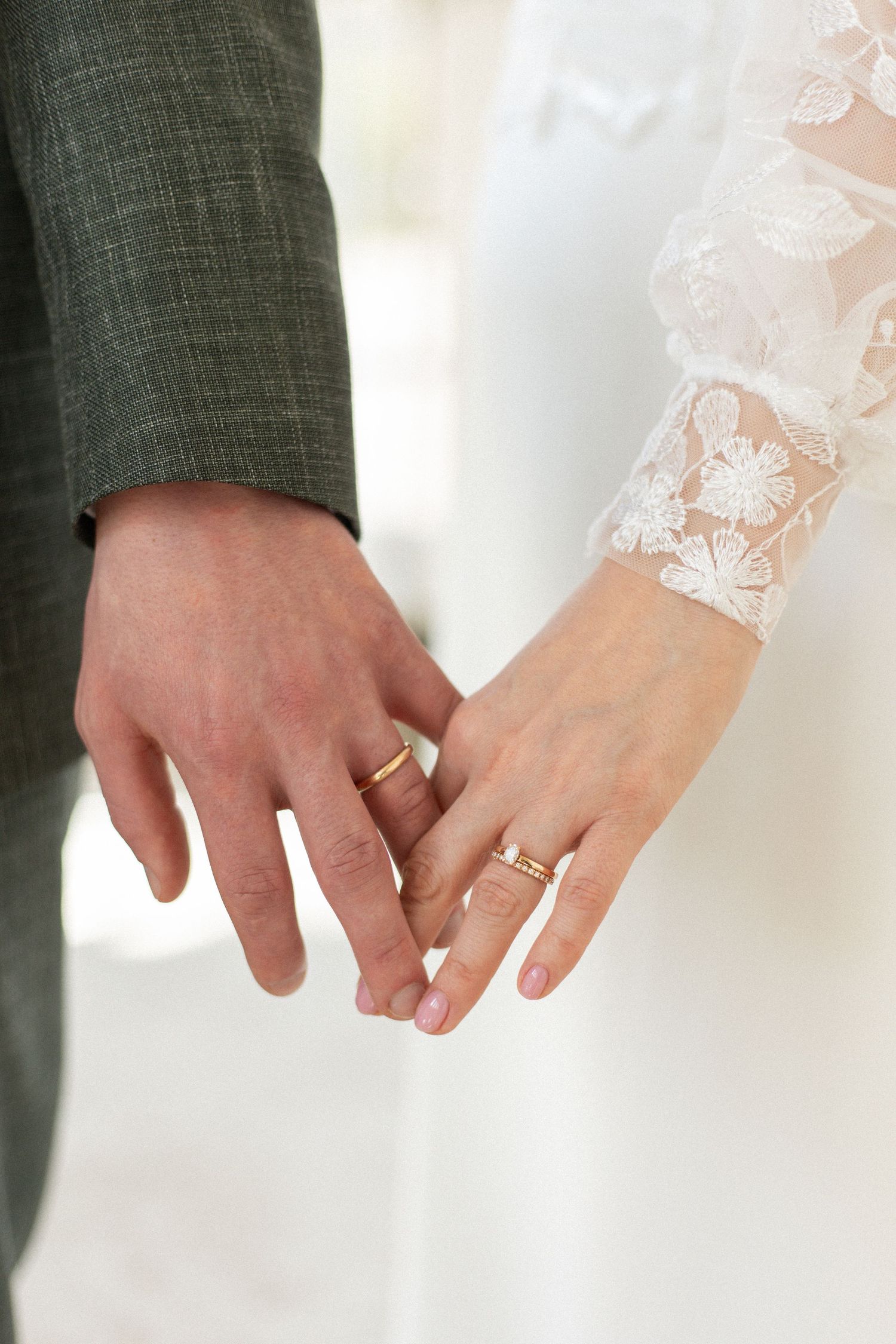 Close-up of newlyweds' hands showing wedding rings against white lace and gray suit fabric.