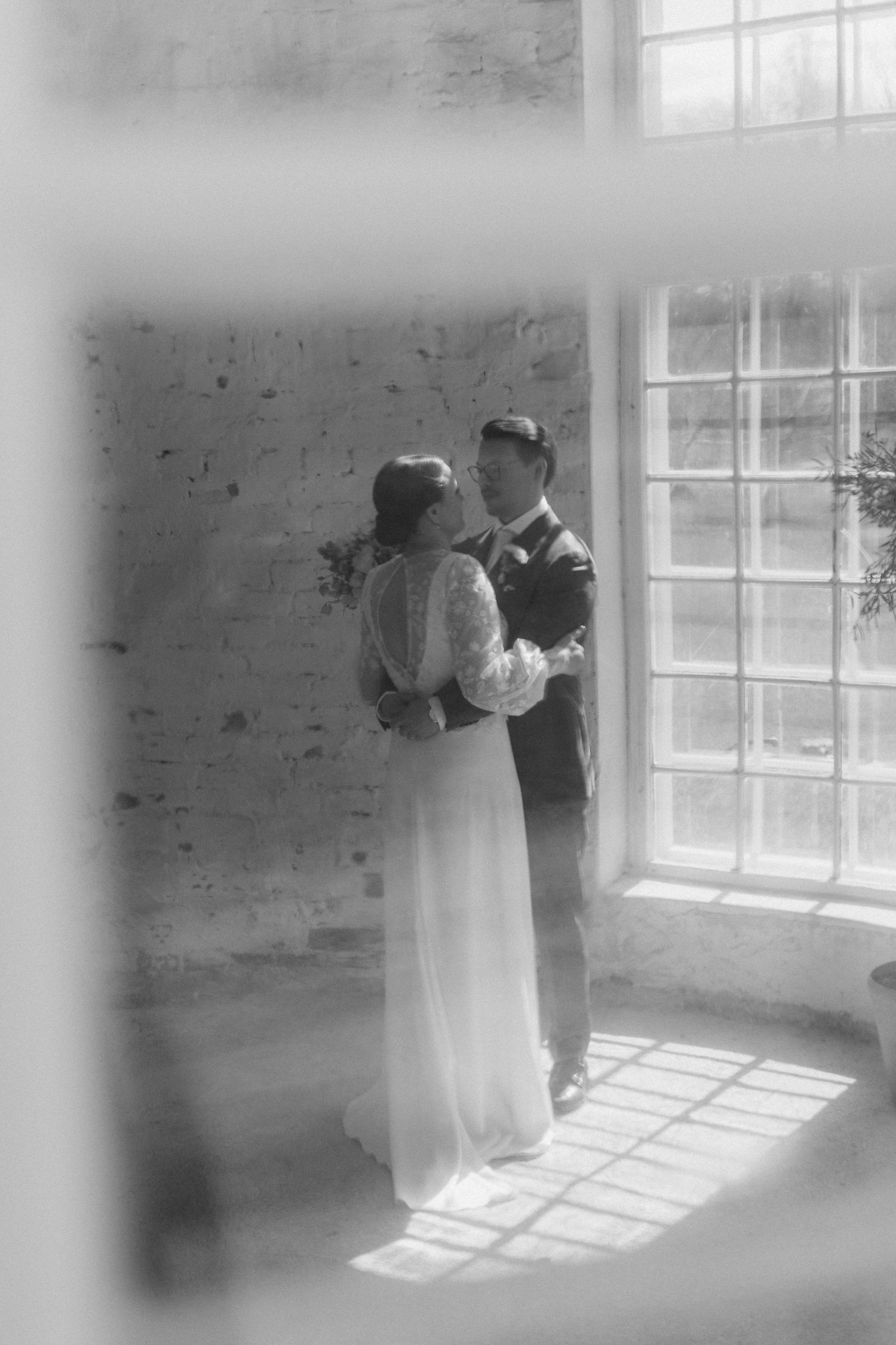 Romantic black and white photo of newlyweds by a sunlit window.