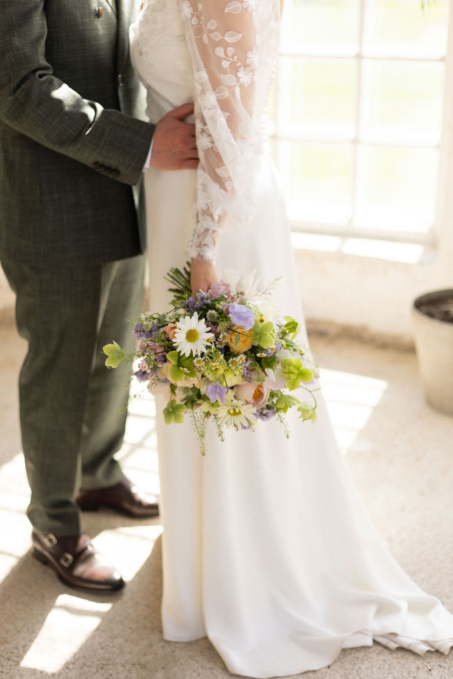Wedding detail shot showing white lace dress against gray suit with spring wildflower bouquet.