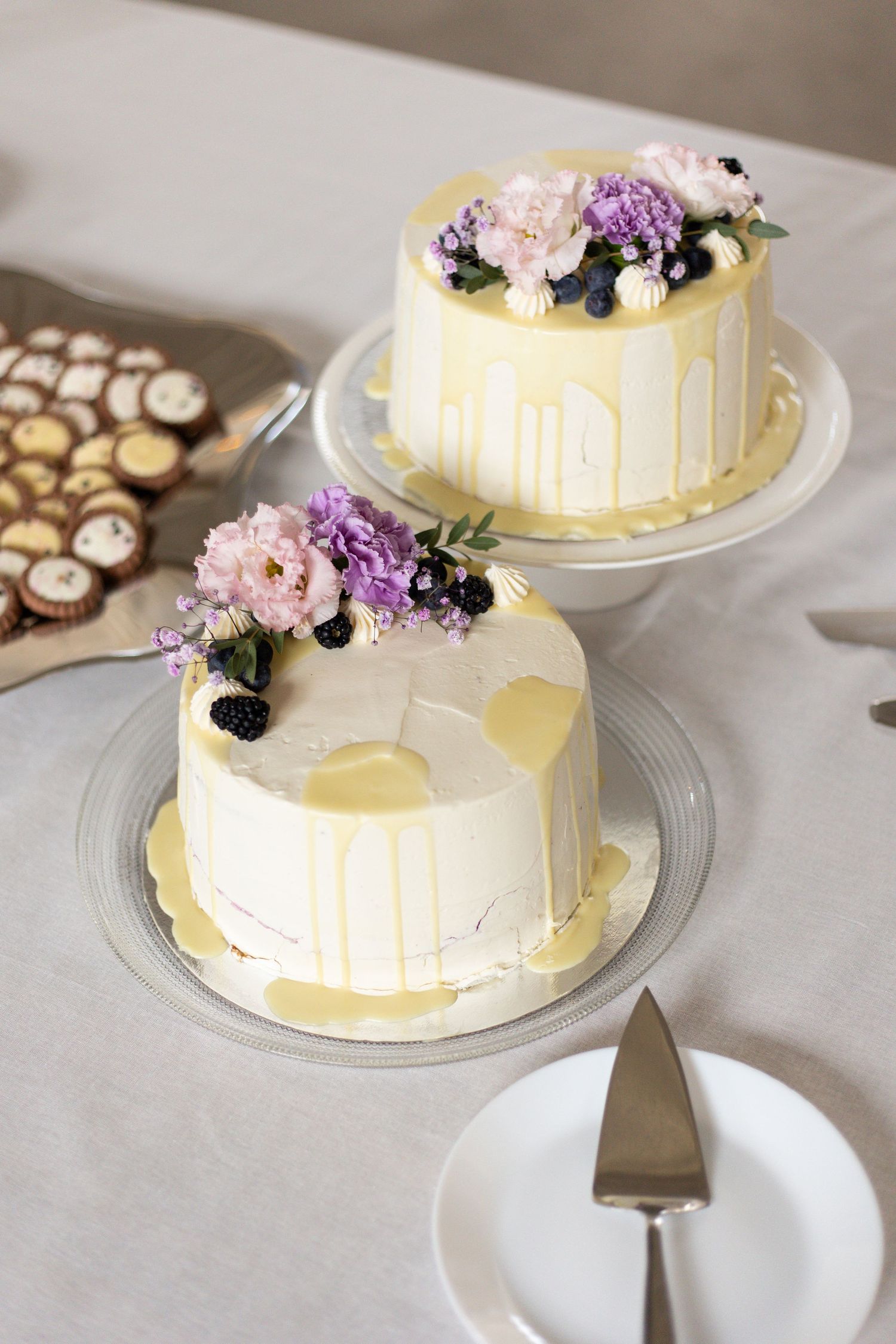 Two elegant white wedding cakes decorated with fresh purple and pink flowers and gold drips displayed on cake stands.