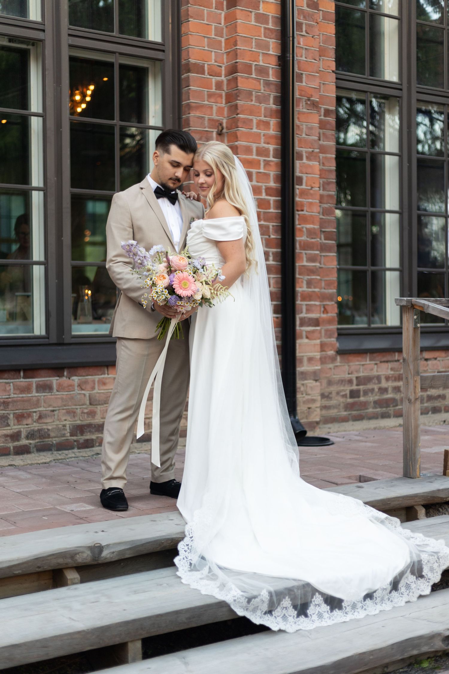 Wedding scene on wooden steps against a brick building with large windows.