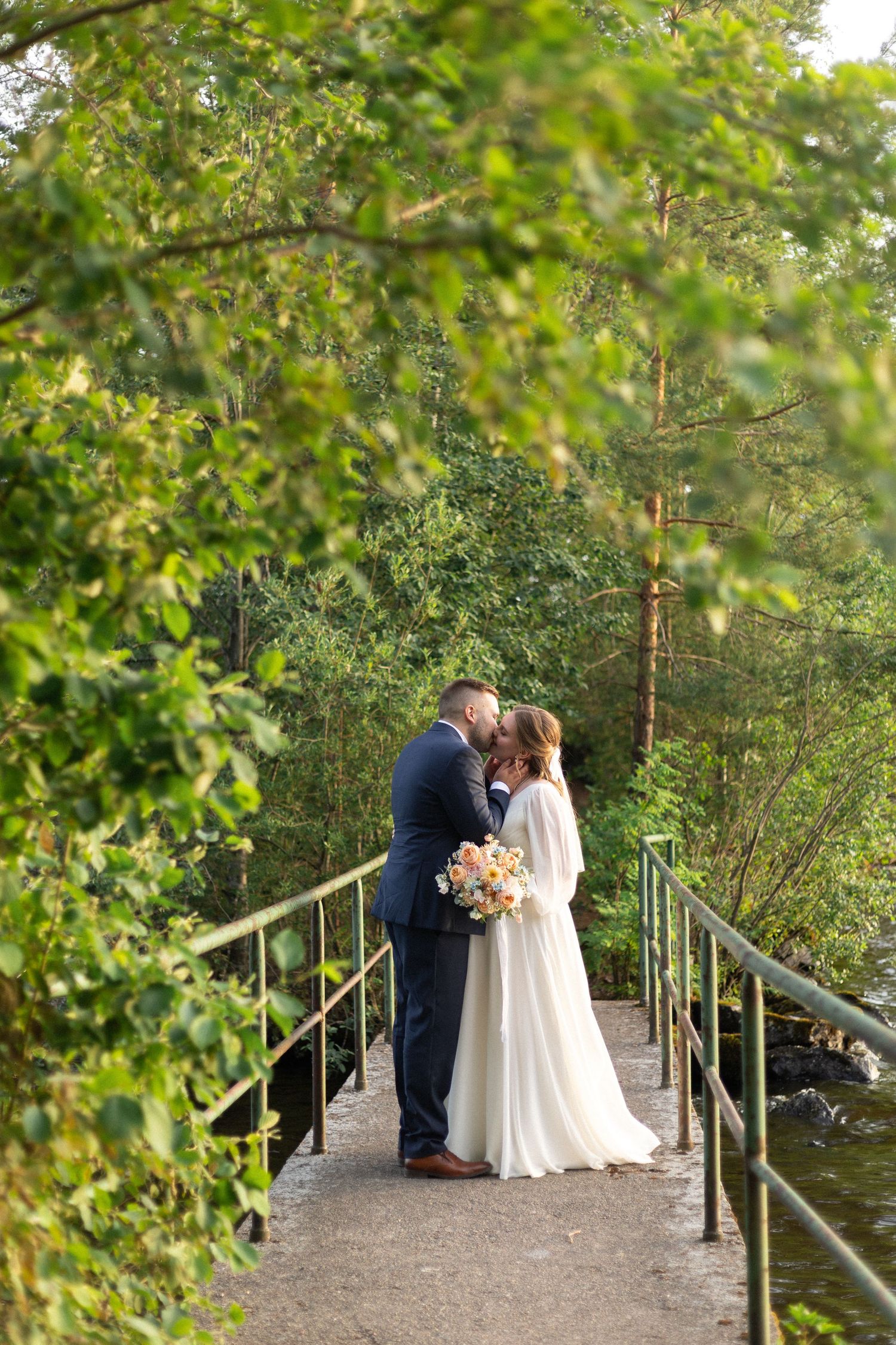 Romantic wedding couple embraces on a rustic bridge surrounded by lush green foliage.