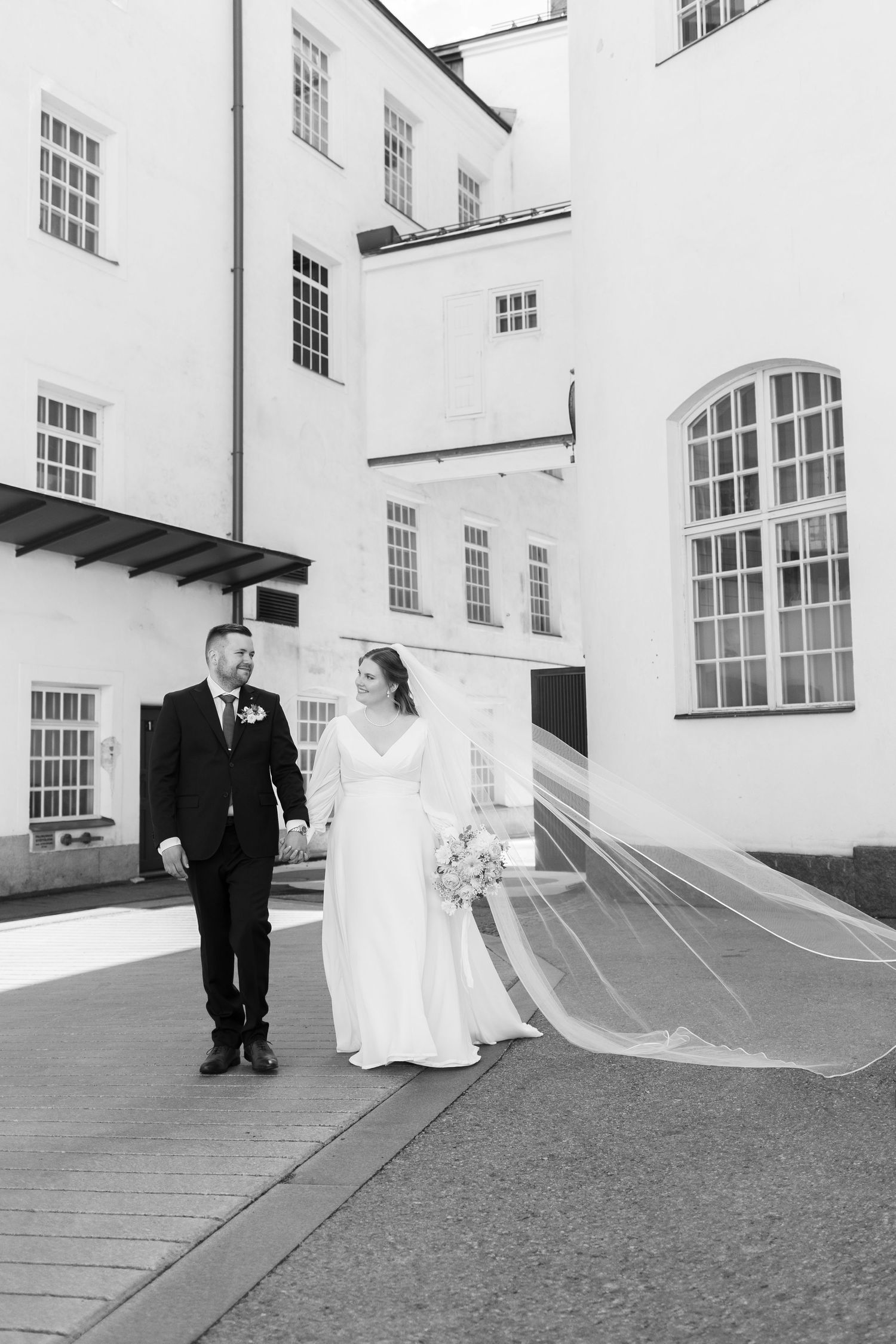 Newlyweds walking together against white building exterior with long flowing veil.