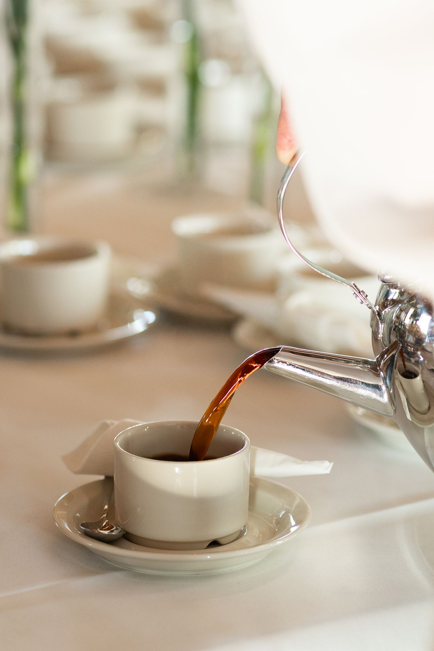 Tea being poured from silver teapot into white china cup on formal table setting.