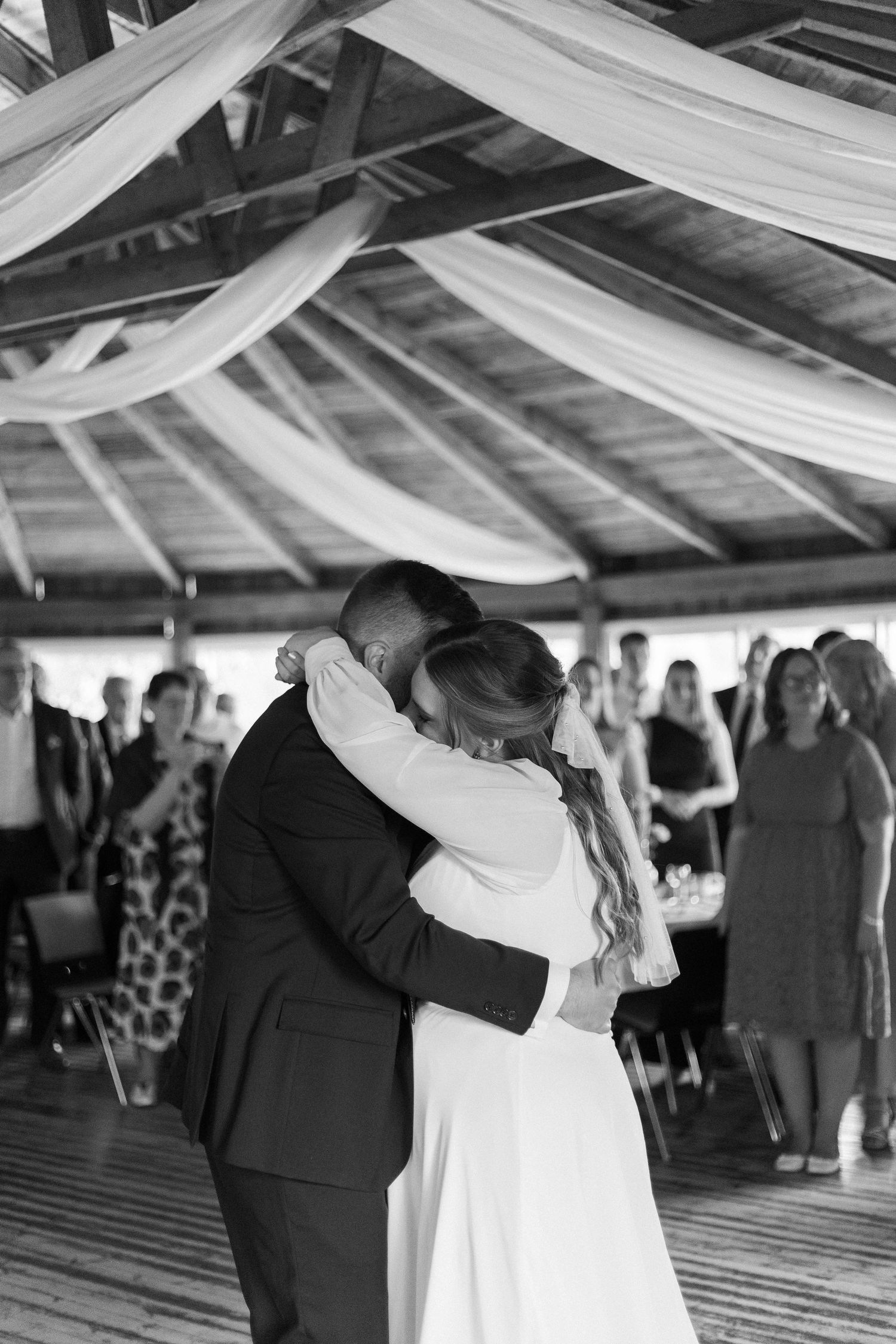 A couple shares their first dance at a wedding reception under draped white fabric decorations while guests look on.