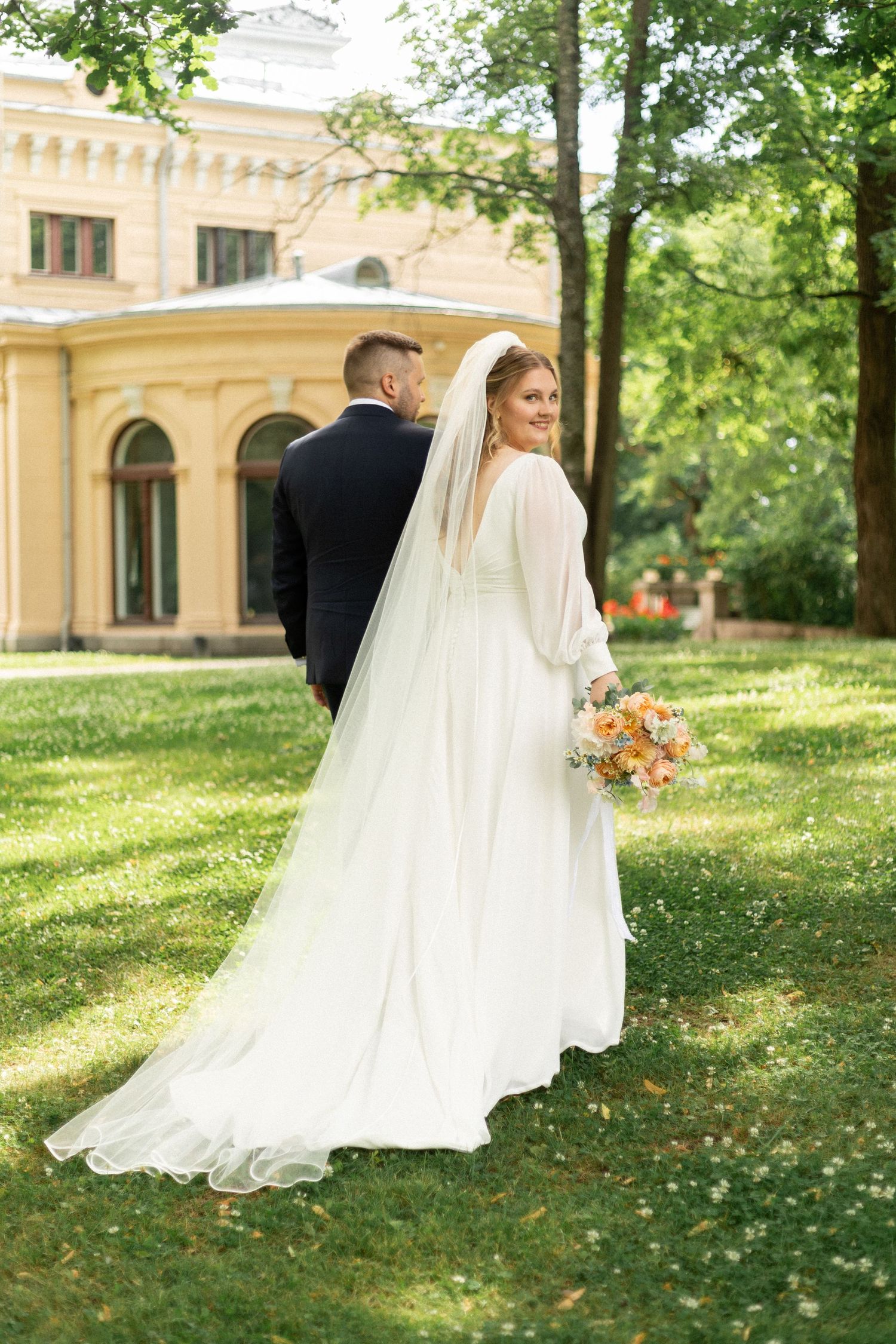 Bride in flowing white gown with long veil poses near historic yellow building.