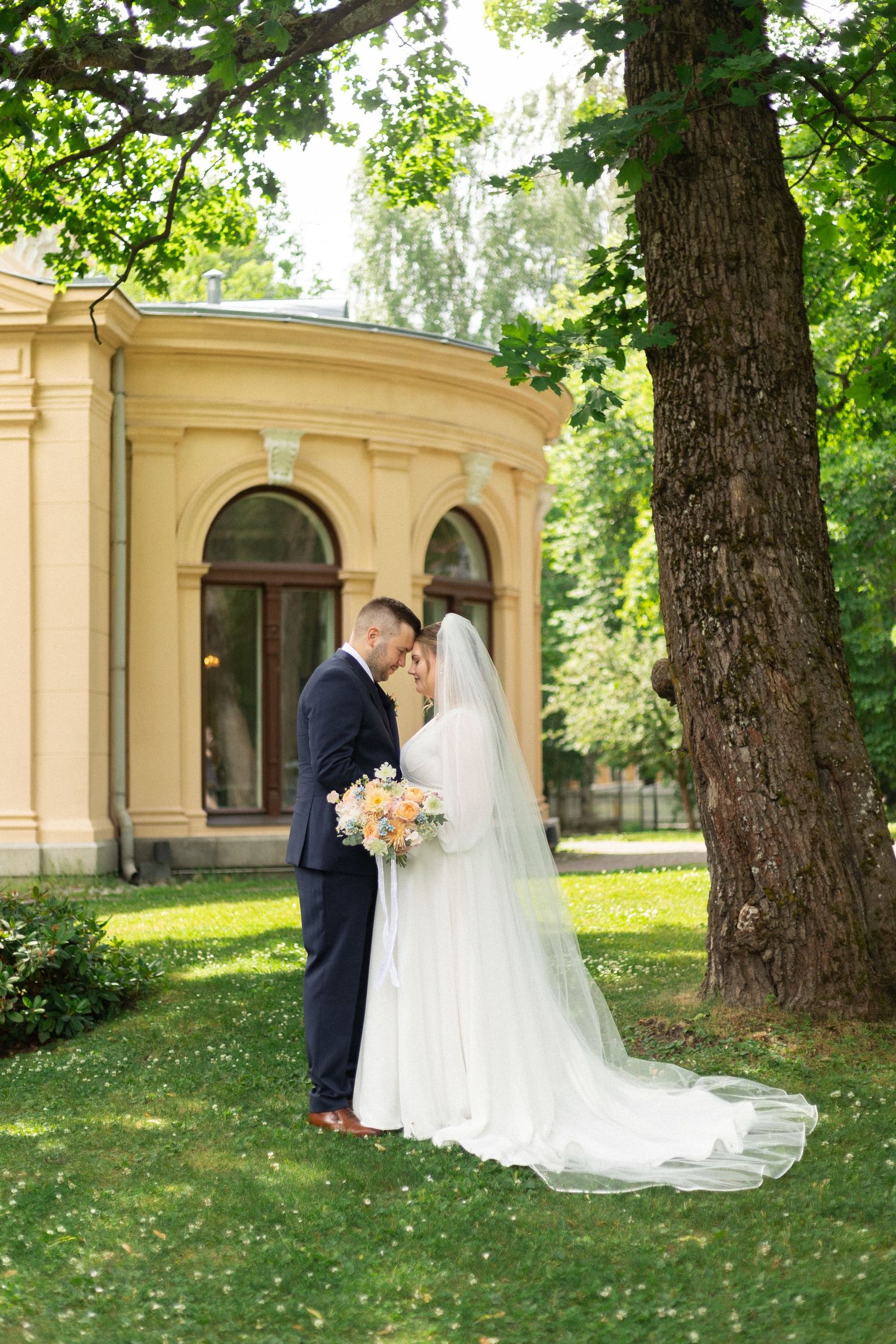 Wedding couple shares intimate moment under large tree by historic yellow building.