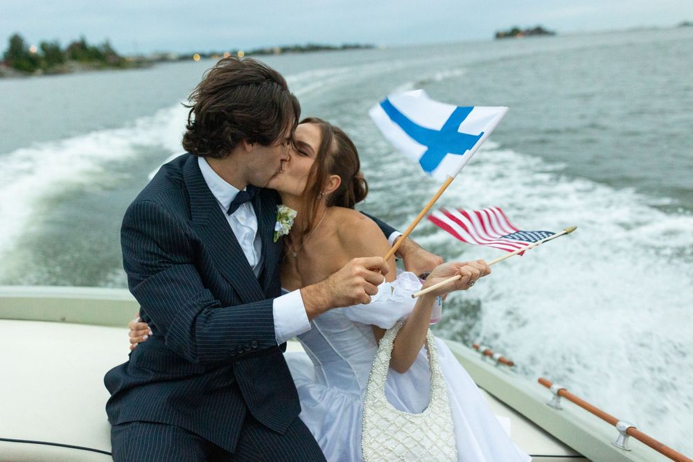 A couple shares a kiss on a boat while holding American and Finnish flags against a splashing ocean backdrop.