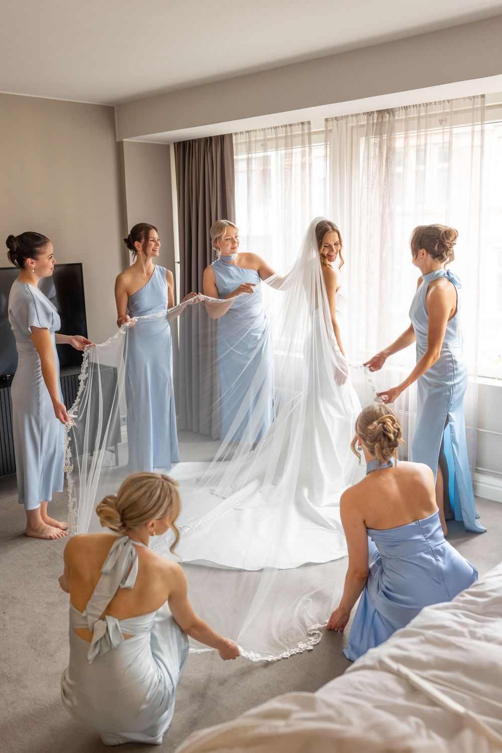 Bridesmaids in light blue dresses help arrange a wedding gown's train in a bright hotel room.