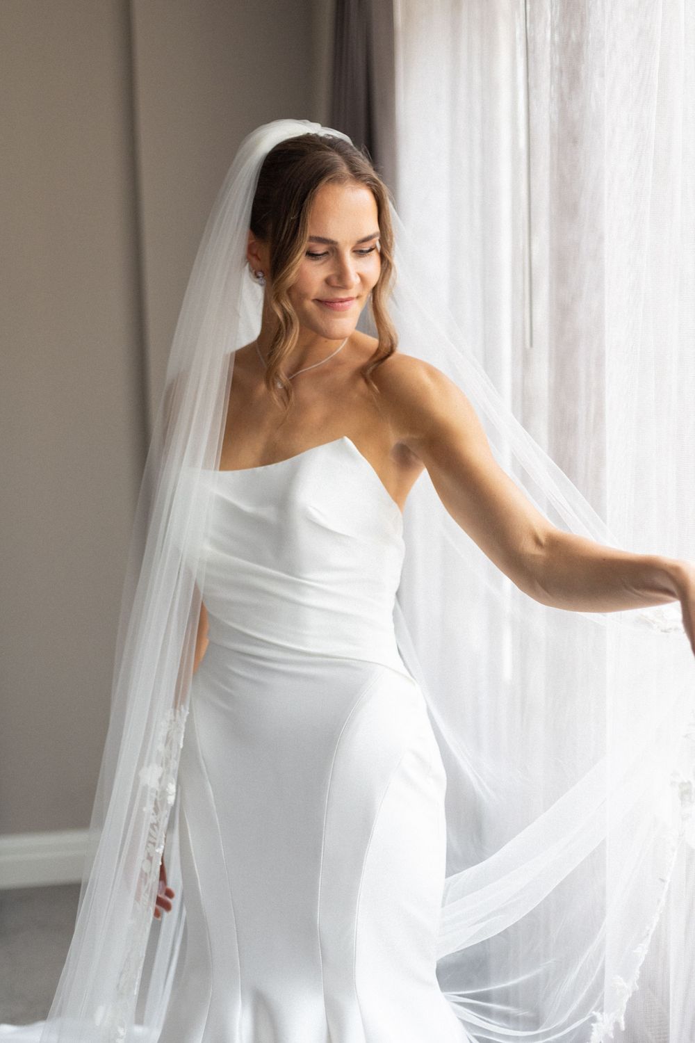 Elegant bride in a sleek white wedding dress and flowing veil poses gracefully near a window in natural light.