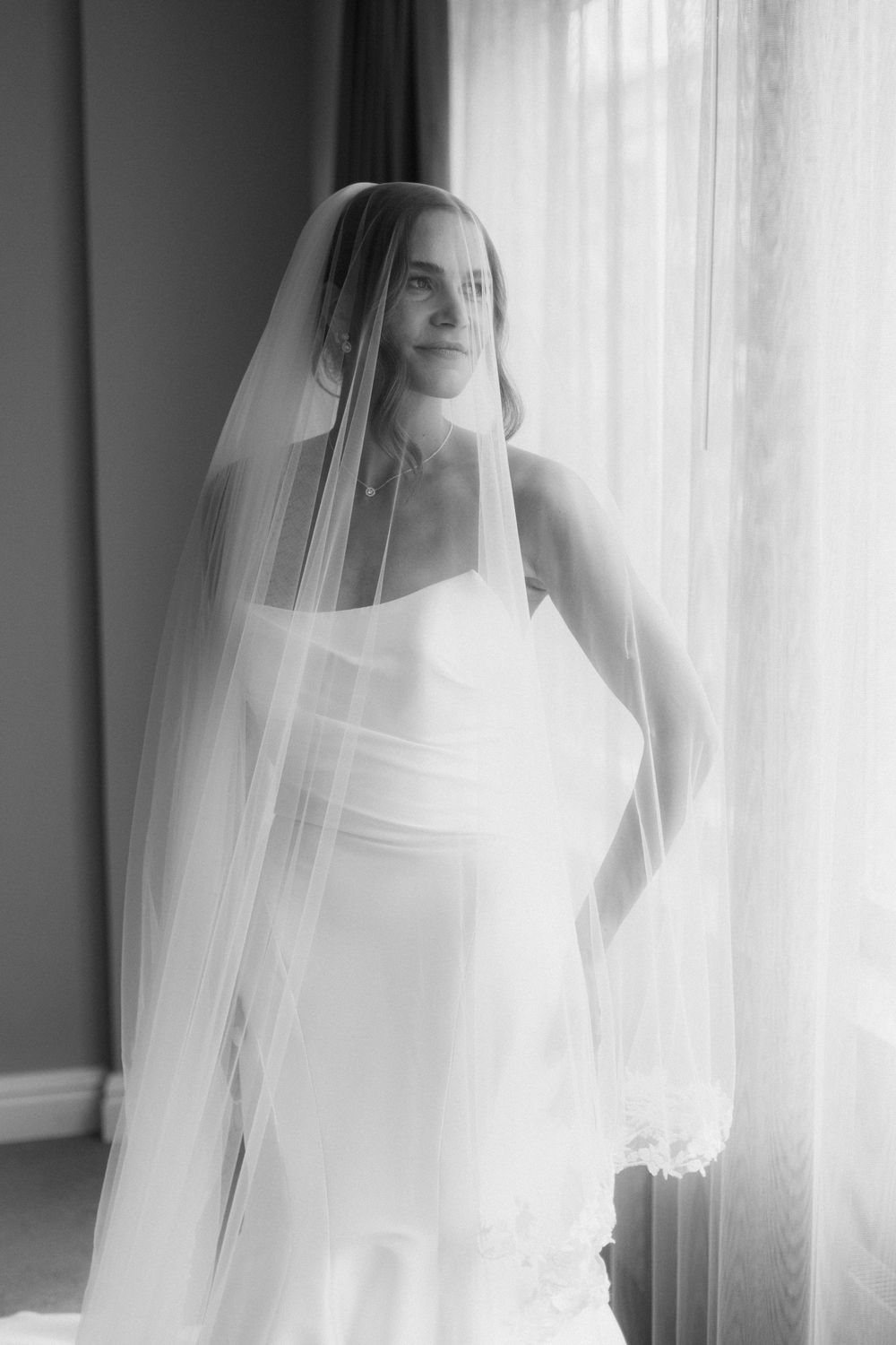 A bride in a white wedding dress and veil poses by a window in a series of ethereal black and white photographs.