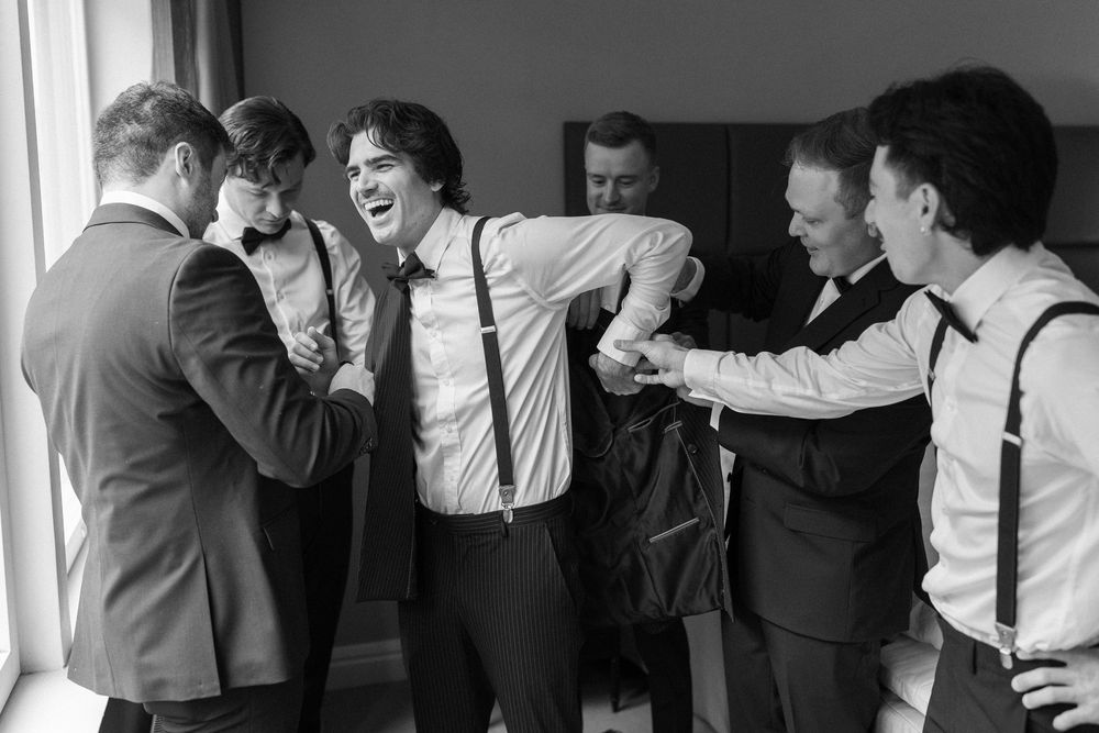 A black and white photograph shows a group of men in formal attire laughing and joking together by a window.