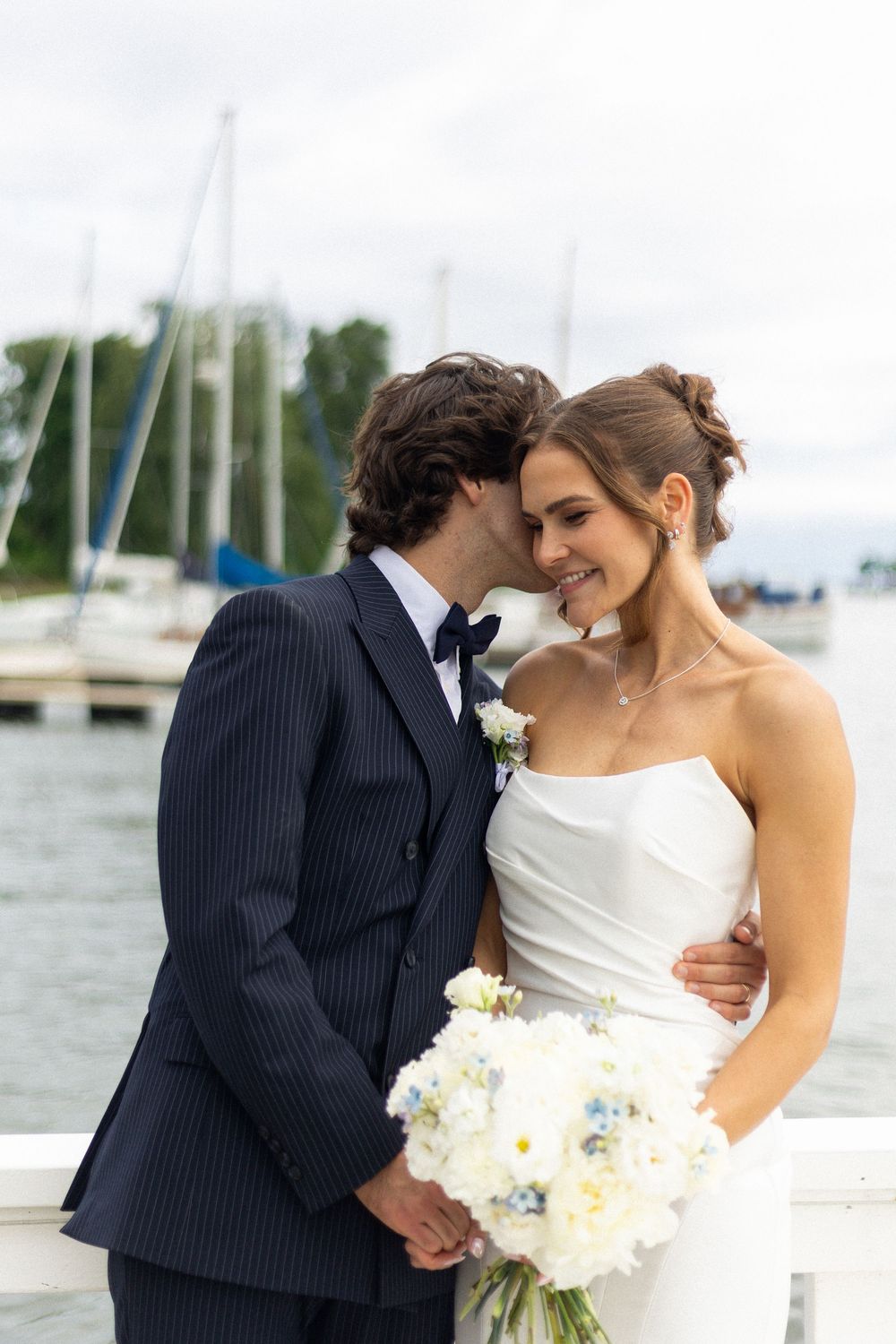 A romantic wedding couple shares an intimate moment by a marina with sailboats in the background.