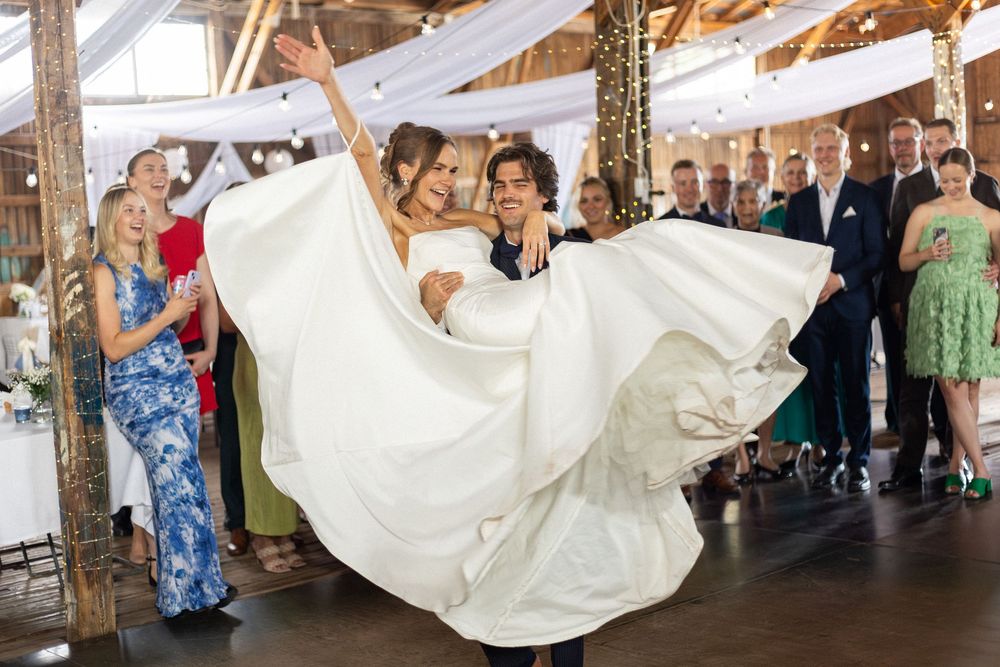Wedding party members lift and swing a bride in her white dress during a lively dance floor celebration.