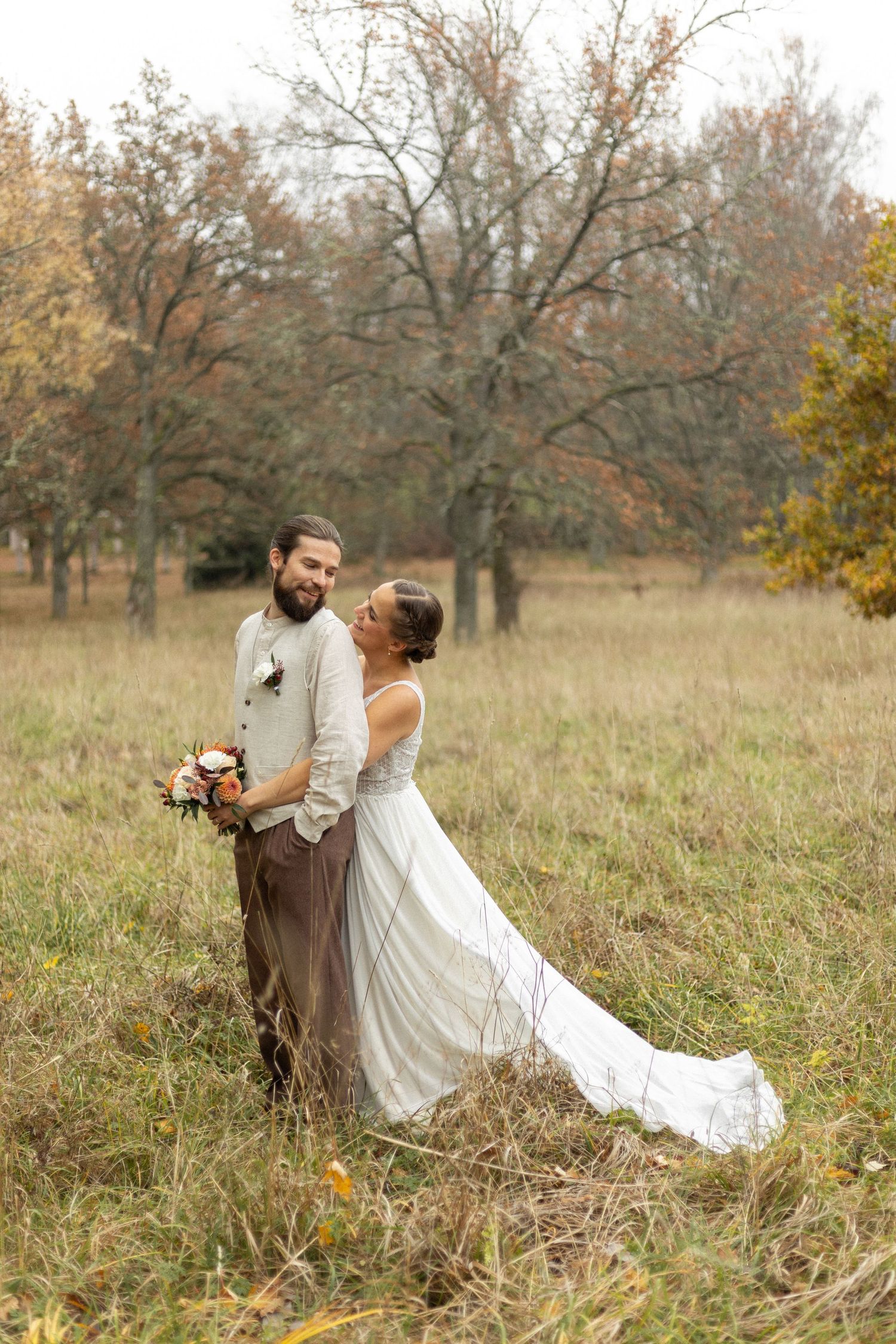 A couple in rustic wedding attire posing in an autumn field with fall foliage.