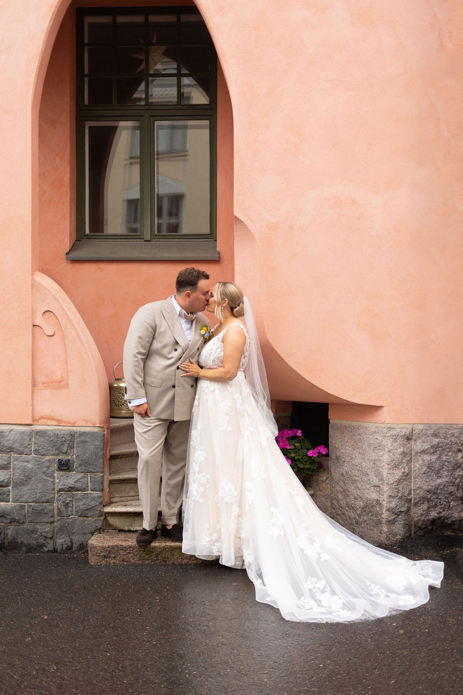 Wedding couple standing in front of pink stucco building with arched windows.