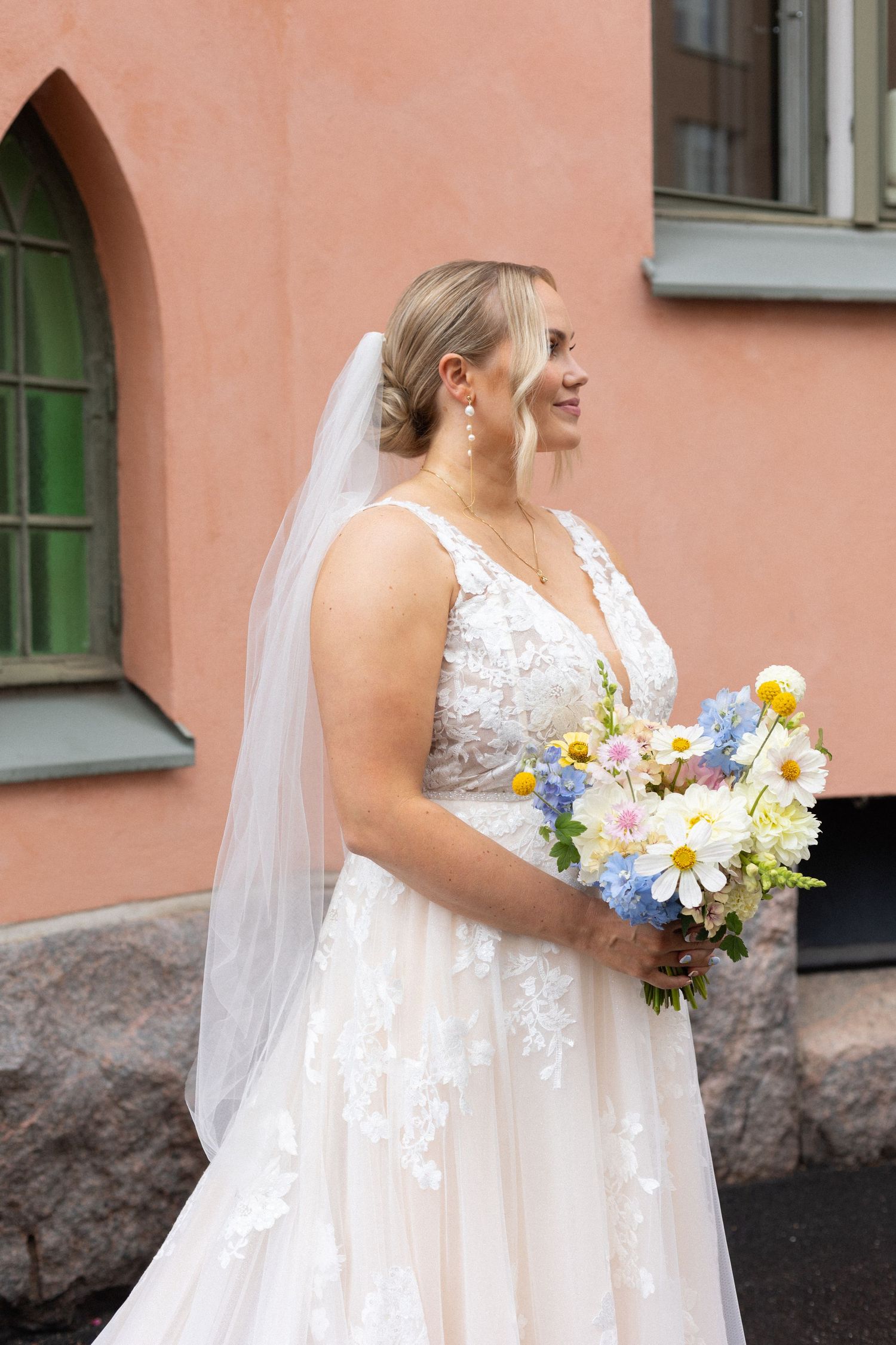 Bride holding colorful summer bouquet in front of pink building.