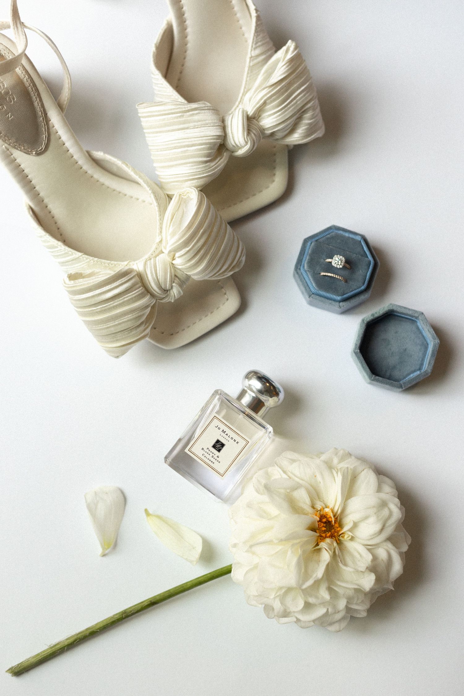 Wedding accessories including white heels, perfume bottle, ring box, and white flower on light background.