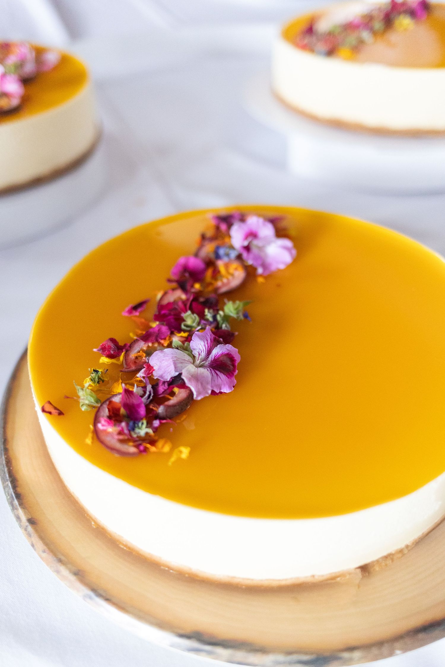 Elegant mango cheesecake topped with purple edible flowers on a wooden base.