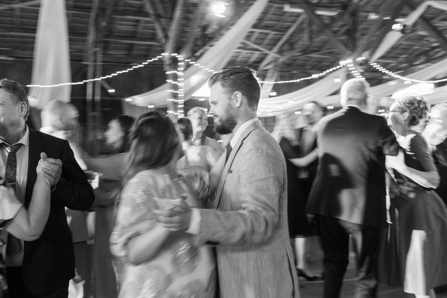 A monochrome image of couples dancing at a reception with string lights overhead.
