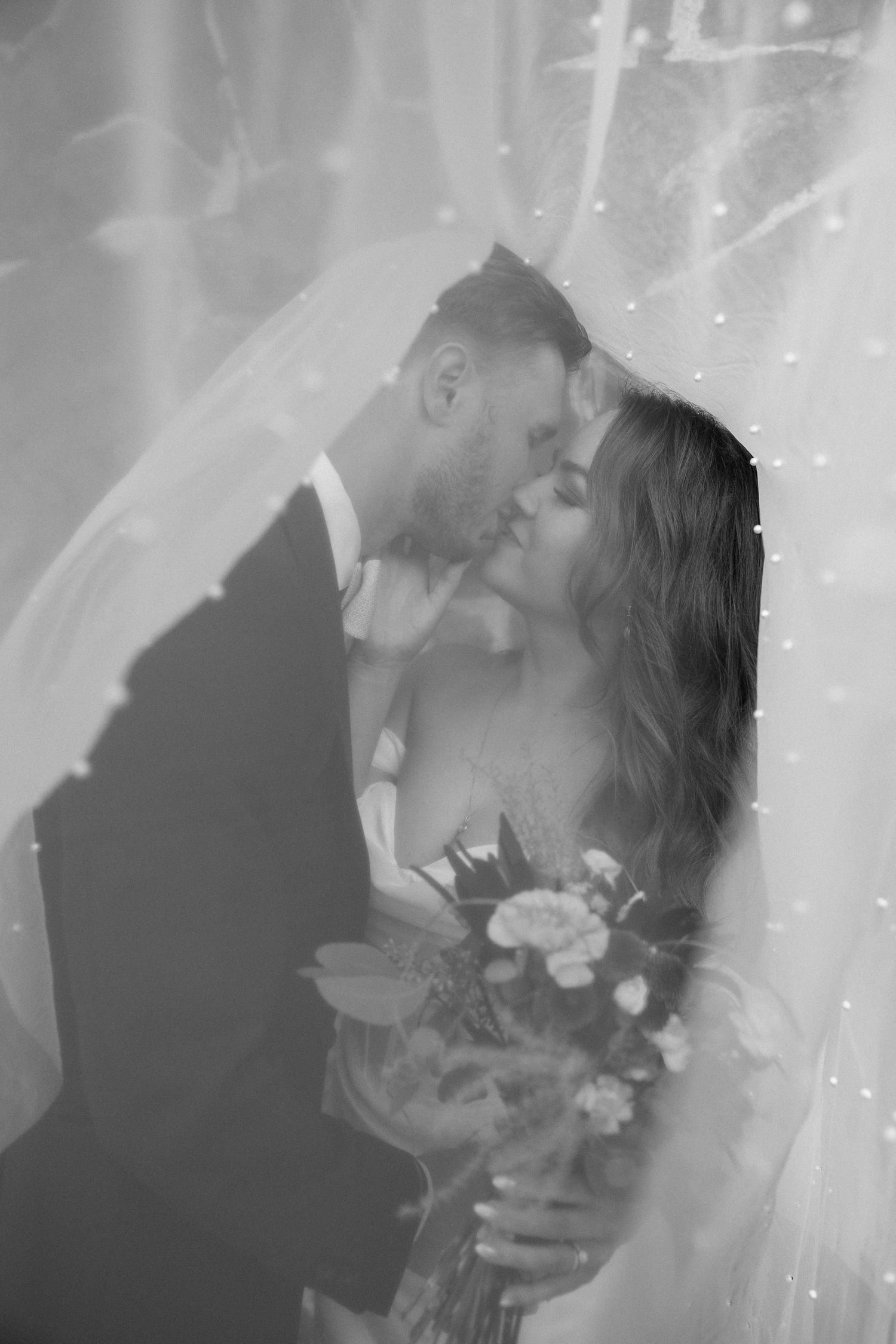 Romantic black and white portrait of a couple under wedding veil with floral bouquet.