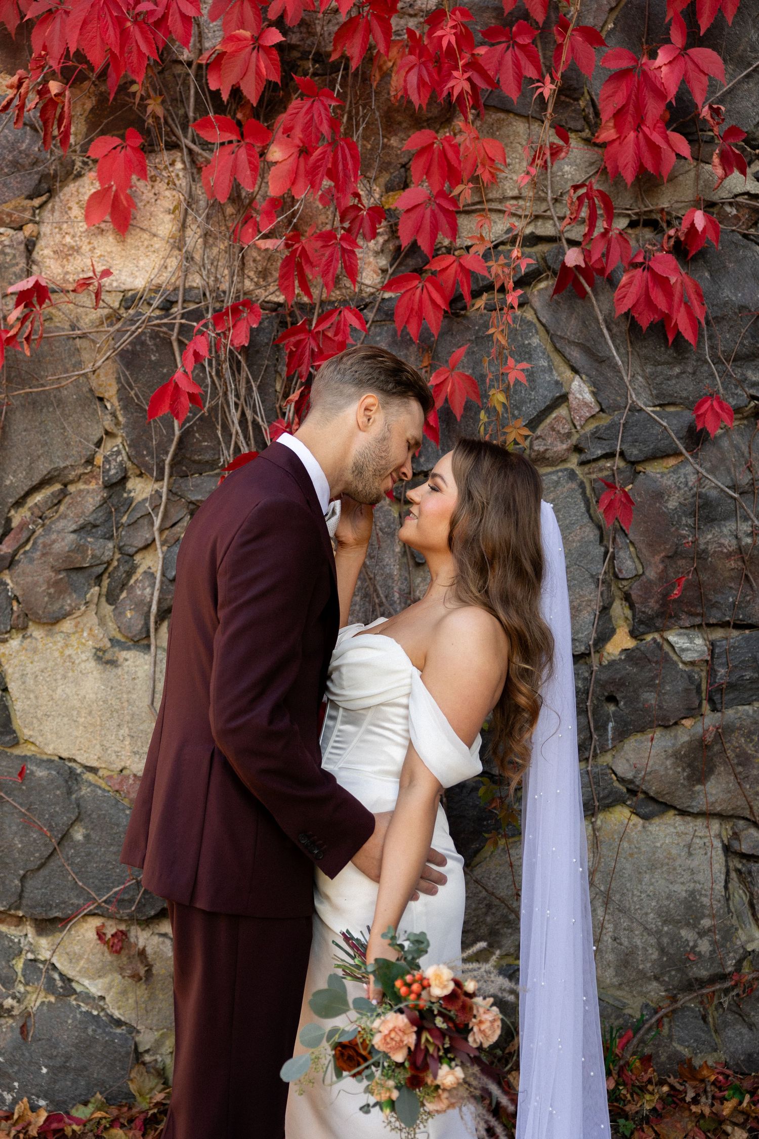 A couple in wedding attire embracing against a stone wall with vibrant red autumn leaves.